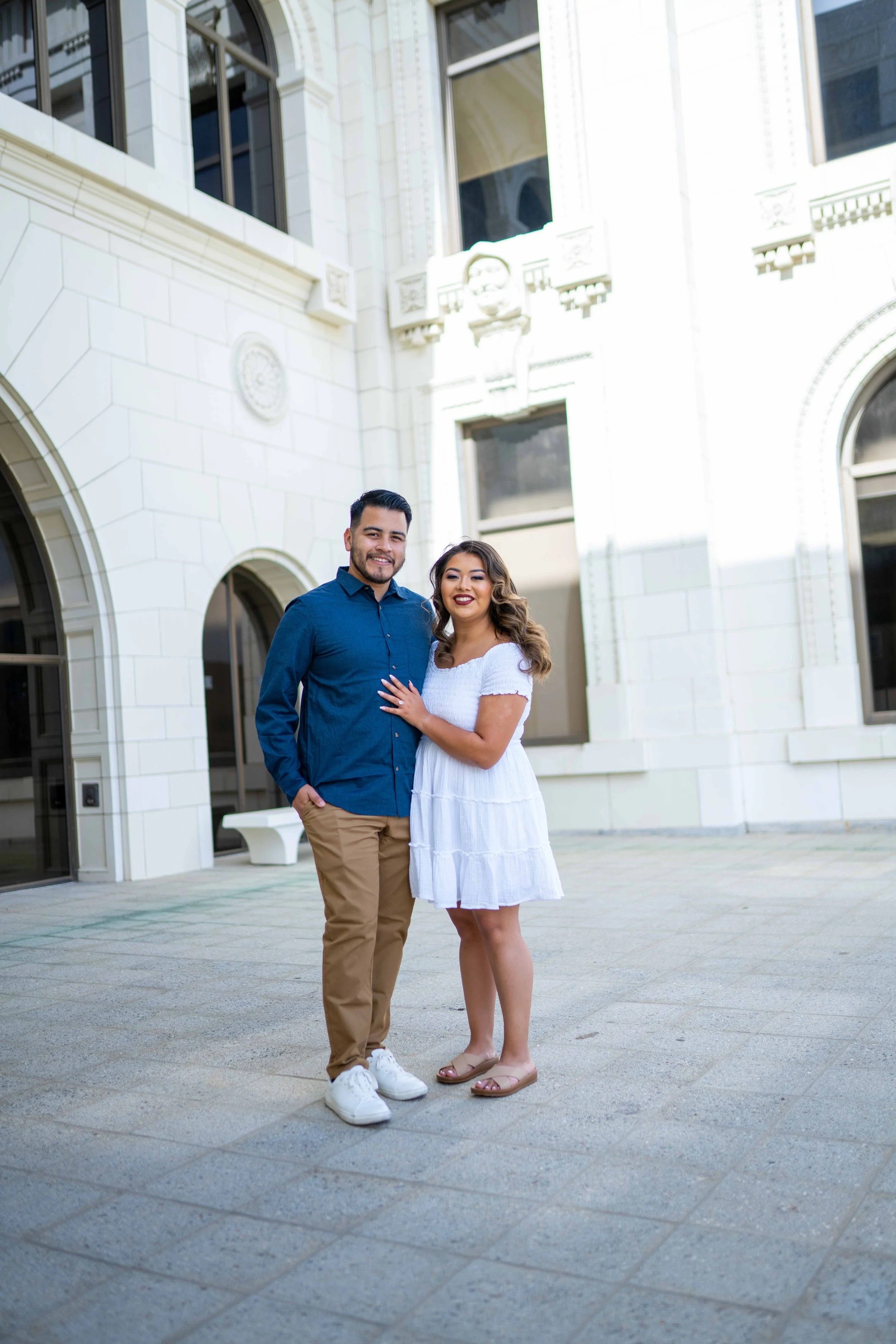A happy couple standing outside in front of a white historic building, smiling at the camera. The man has dark hair and a beard, wearing a blue shirt and khaki pants. The woman has long, wavy hair, wearing a white dress and sandals.