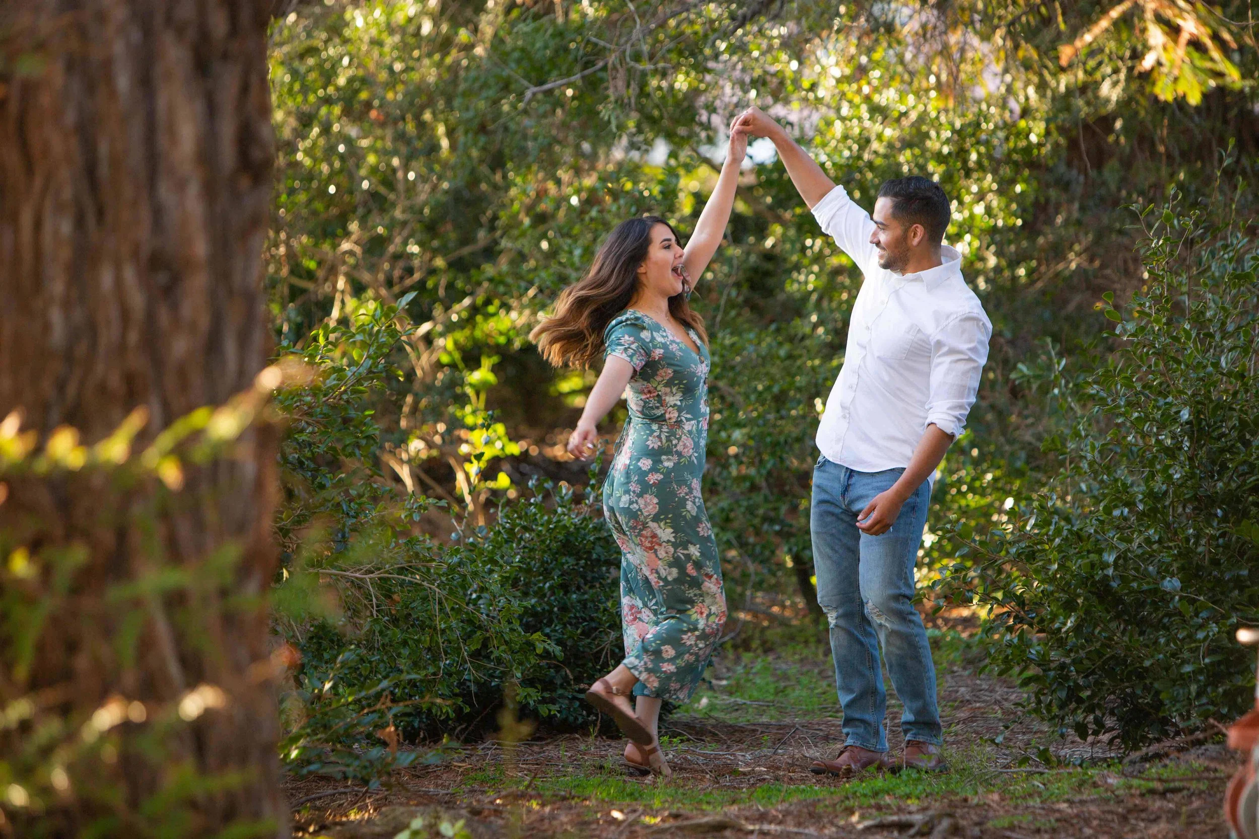 A couple dancing and enjoying outdoors in a forested area during daylight, with green trees and bushes in the background.
