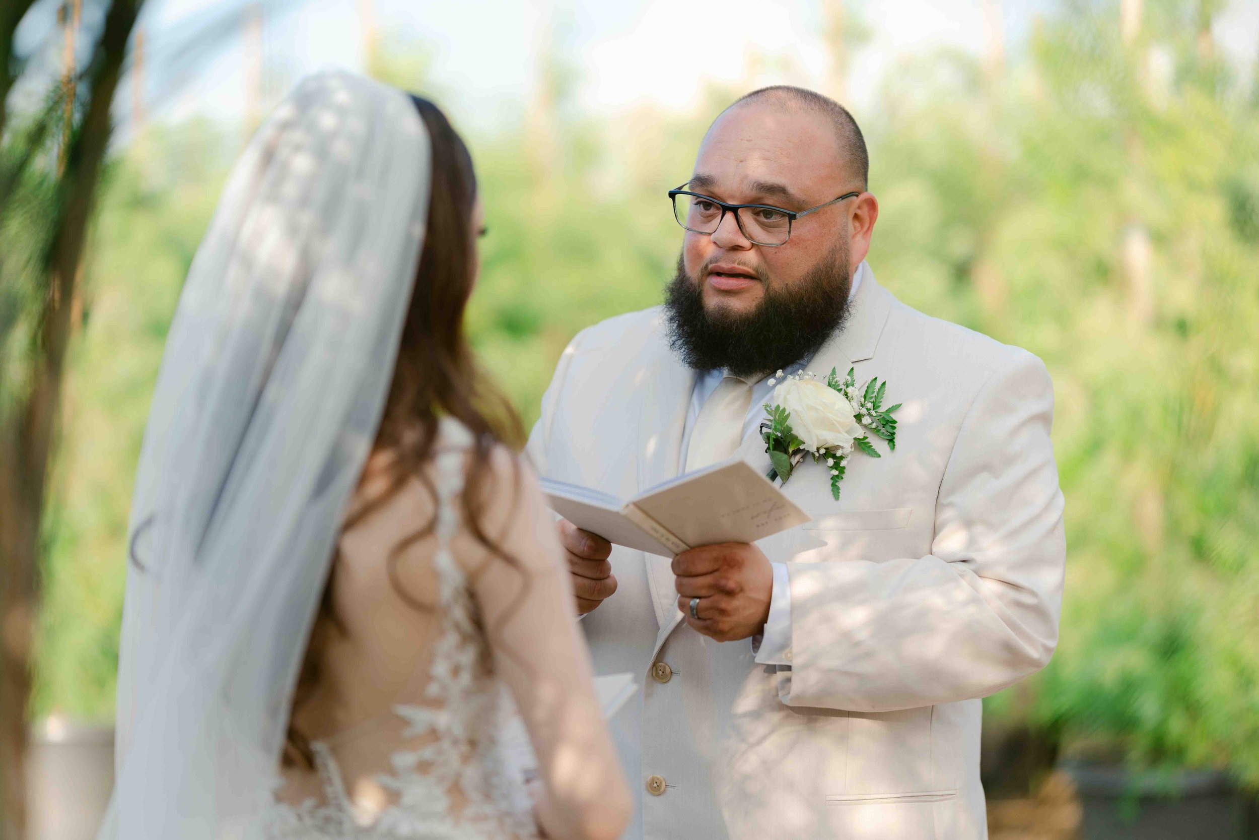 A man in a white suit with a boutonniere and glasses officiates a wedding ceremony outdoors, reading from a book to a woman in a wedding dress with a veil, with greenery in the background.