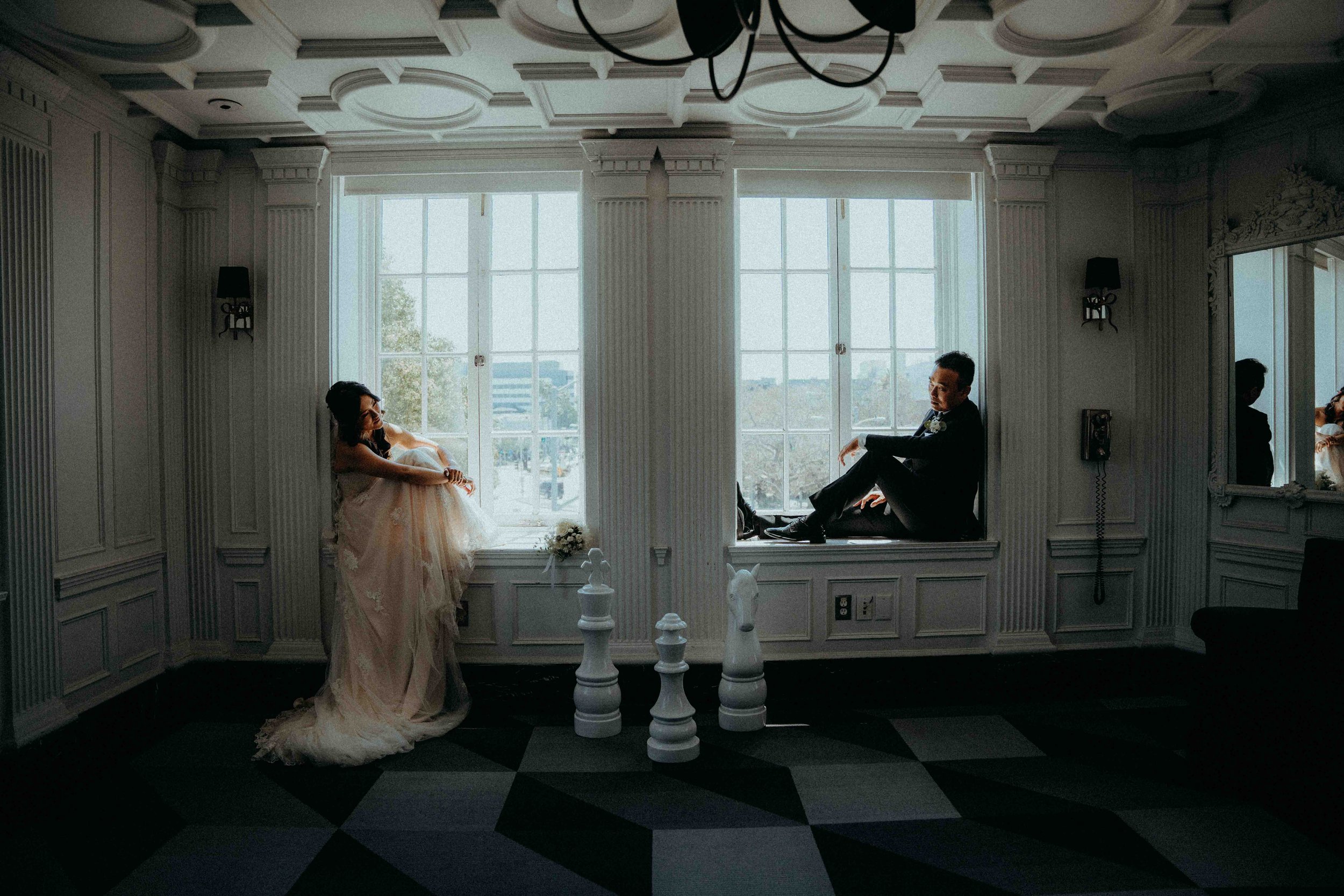 A woman in a wedding dress sitting on a window sill on the left, and a man in a suit sitting on a window sill on the right, in a room with ornate white paneling and large windows.