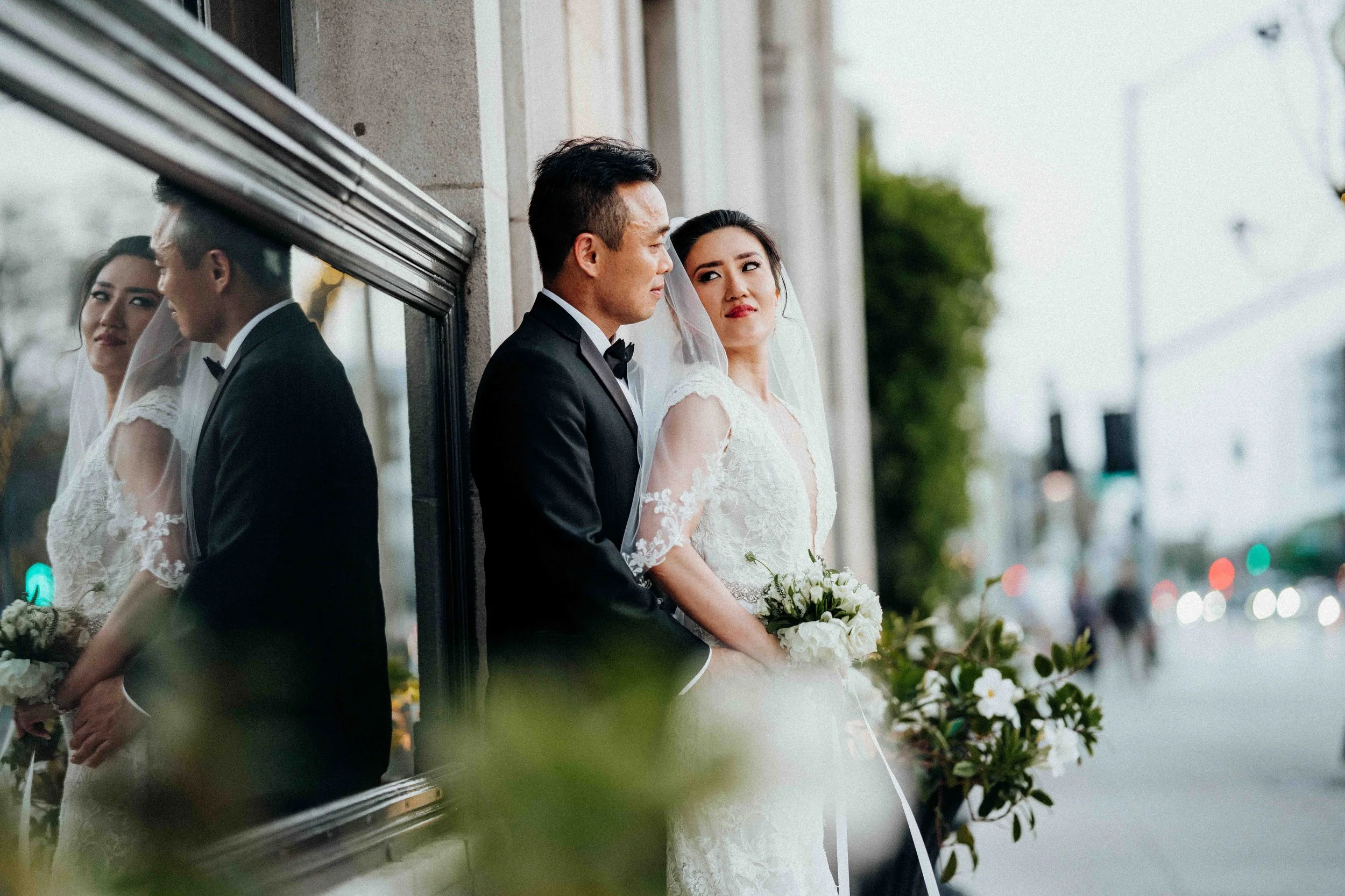 A bride and groom in wedding attire standing outdoors near a glass window, with reflections of themselves. The bride is holding a bouquet of white flowers and looking at the groom, who is dressed in a black tuxedo and looking at the bride. The backgr