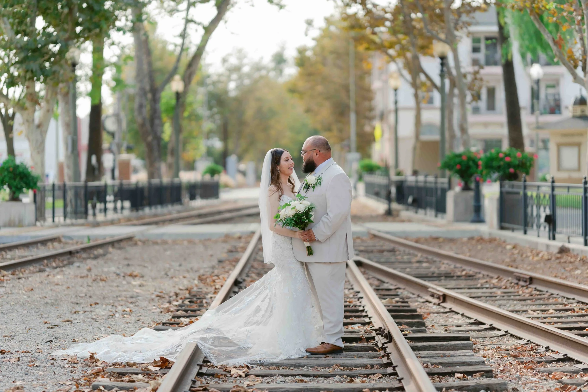A bride and groom in wedding attire standing on tram tracks holding hands and smiling at each other.