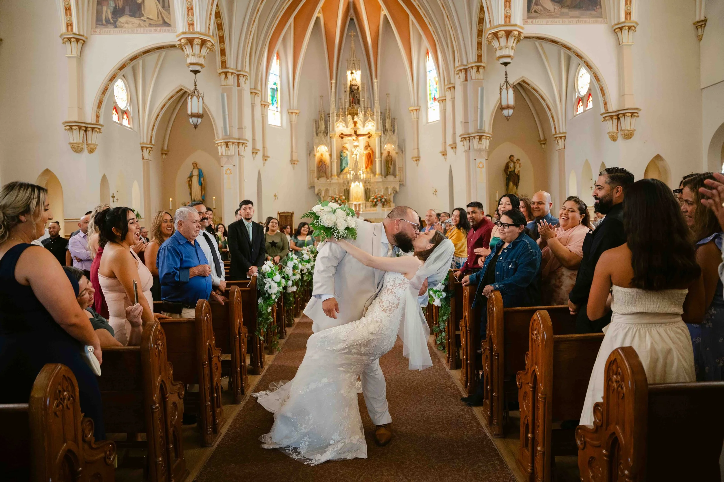 A newlywed couple sharing a kiss at the altar in a church, surrounded by cheering guests and stained glass windows.