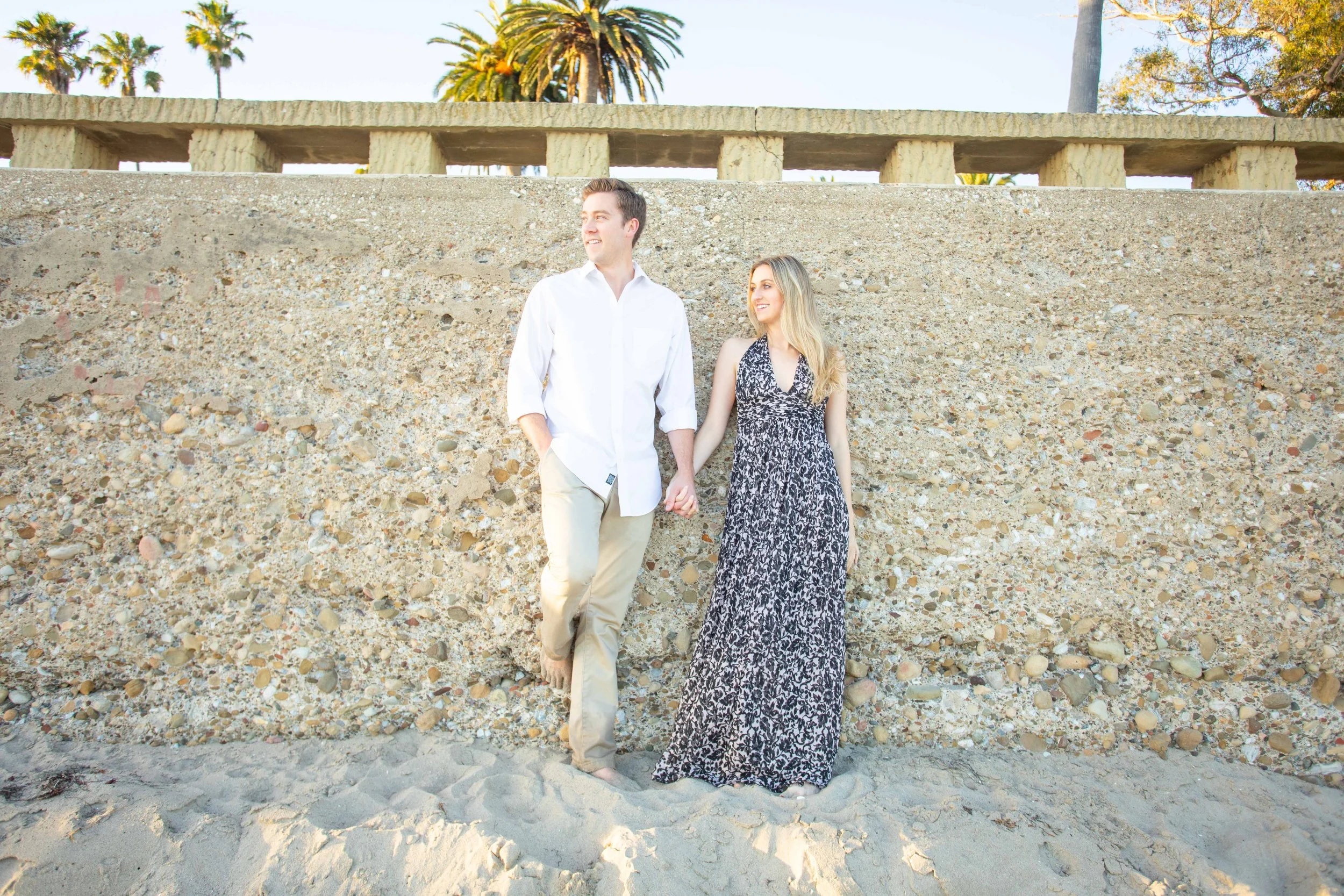 A young couple holding hands while standing barefoot on the sand at the beach, leaning against a concrete wall with palm trees and a clear sky in the background.