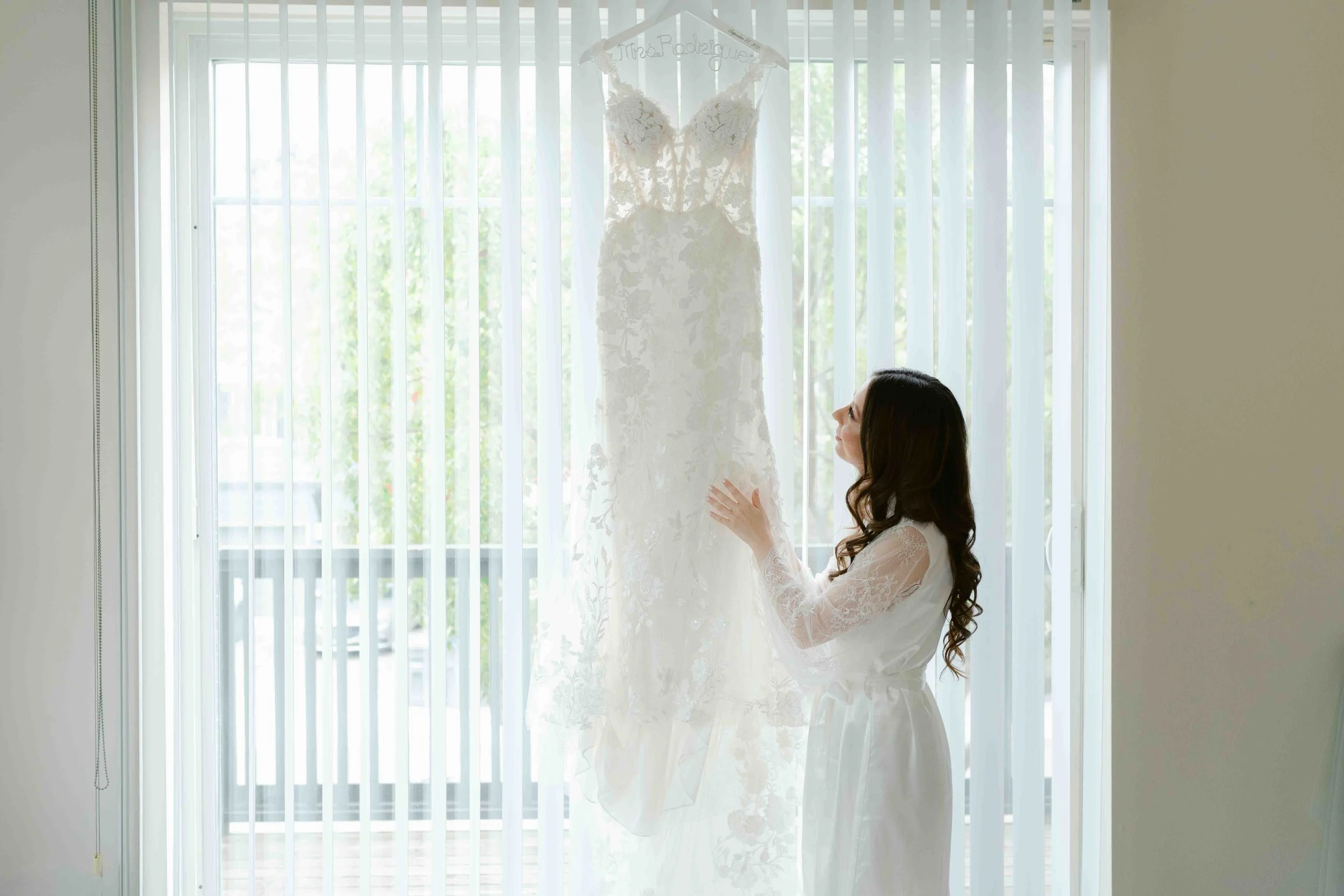 A woman in a white robe looking at a wedding dress hanging in front of window blinds.
