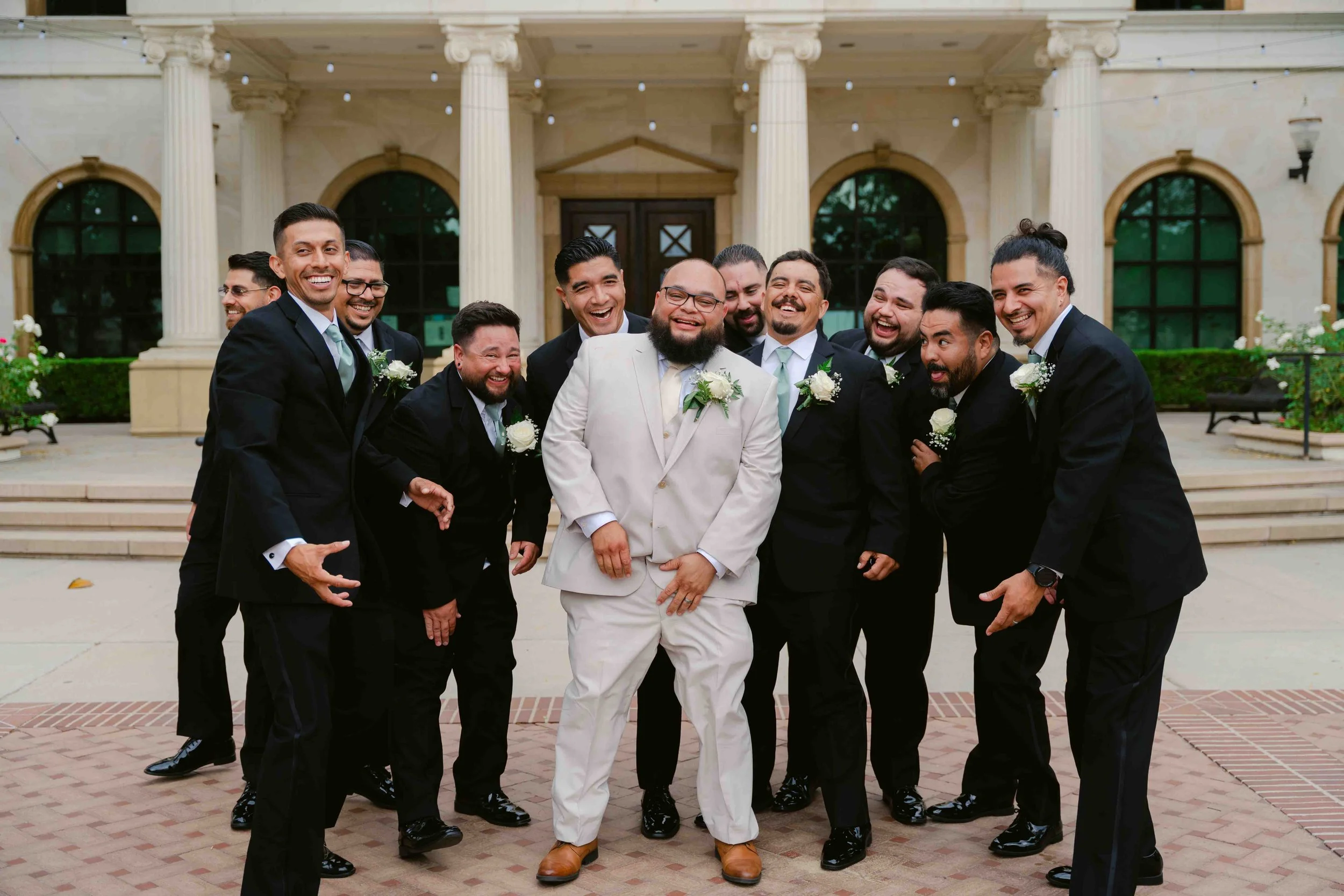 A group of men dressed in formal suits, with boutonnieres, standing happily in front of a building with columns, posing for a photo at a wedding celebration.
