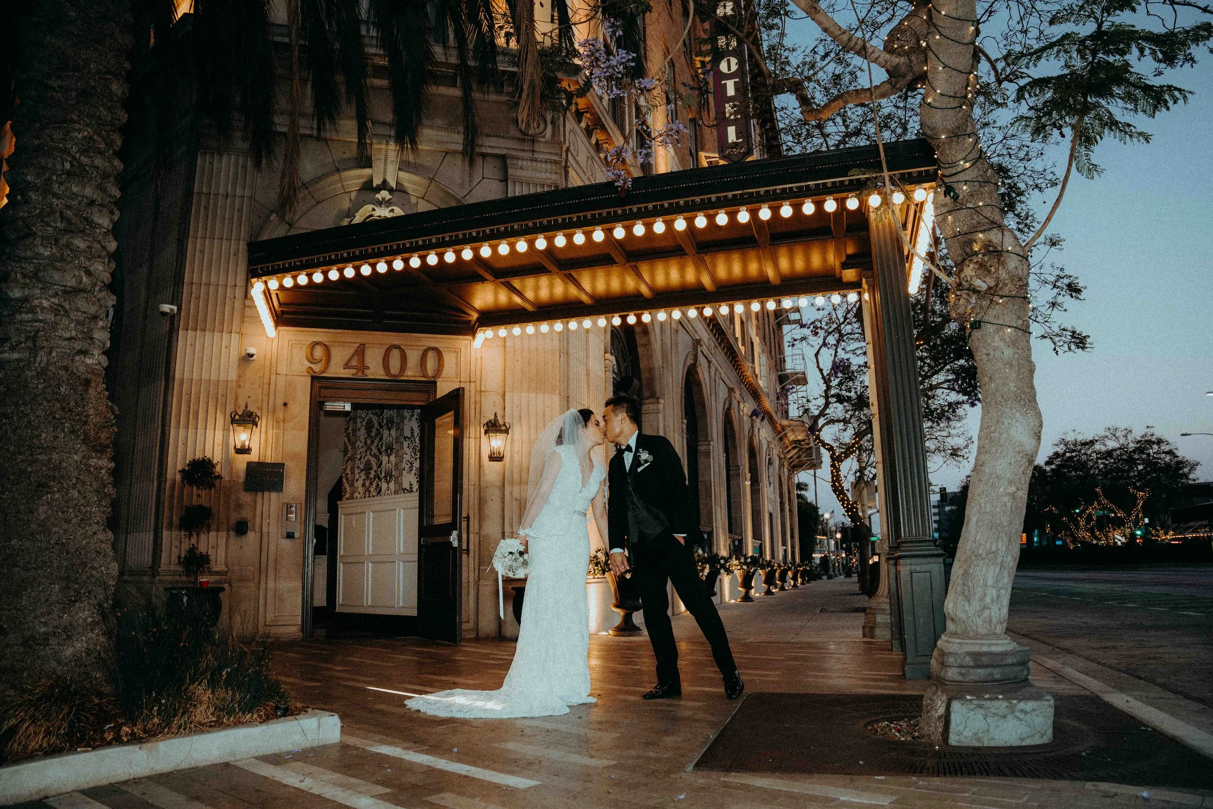 A bride and groom sharing a kiss outside a building with string lights overhead during dusk.
