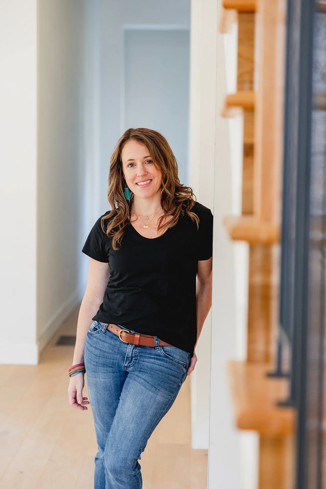 A woman with wavy brown hair, wearing a black t-shirt and blue jeans, standing in a bright corridor with a smile.