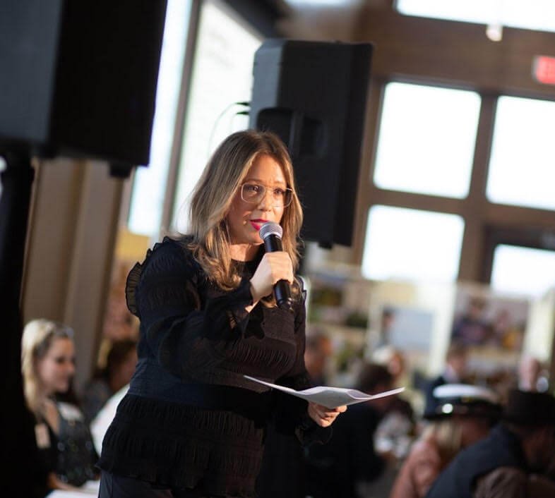 A woman with glasses and shoulder-length hair speaking into a microphone at an indoor event, holding papers in her hand.