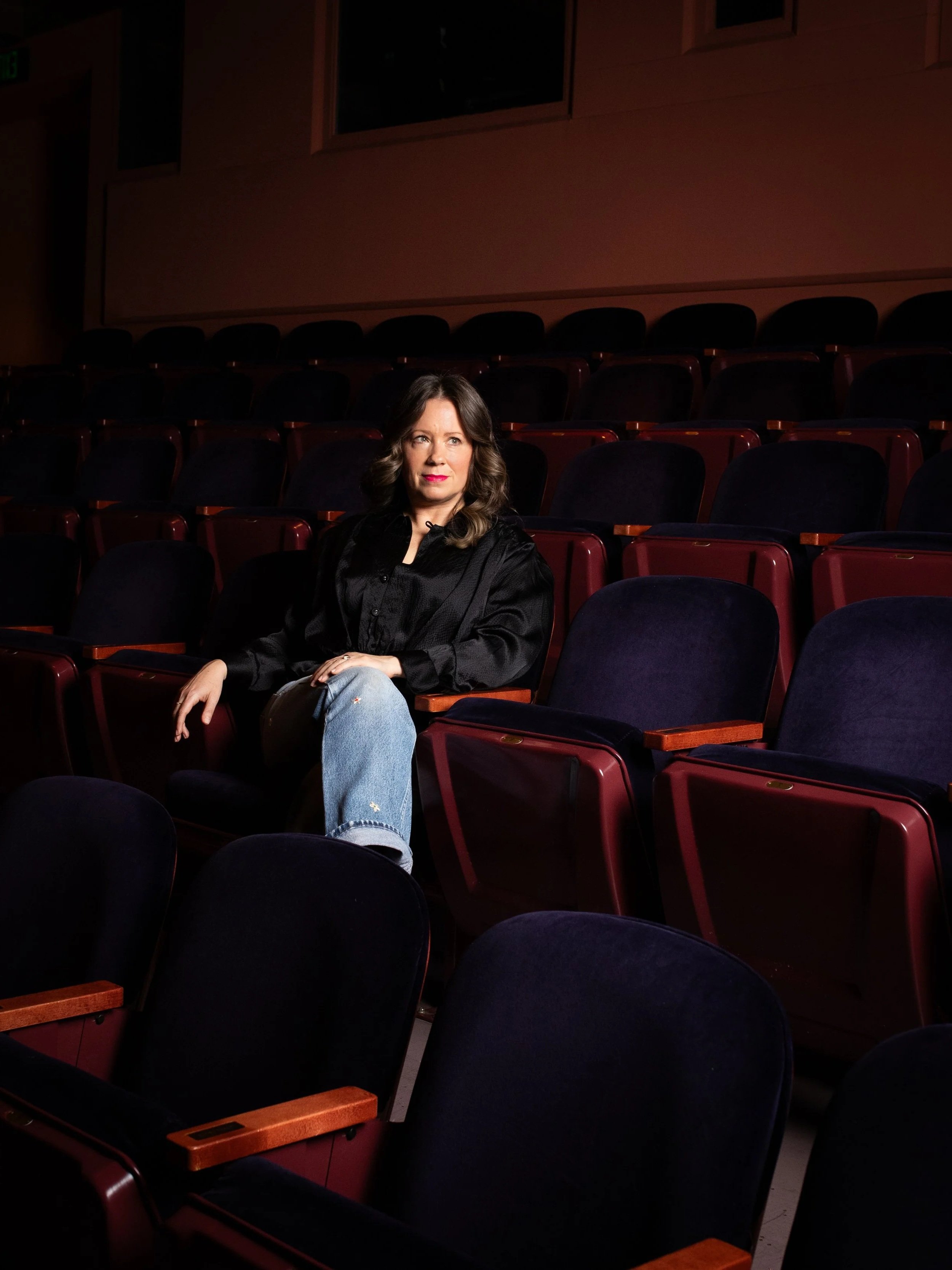 A woman sitting alone in an empty theater with red seats, wearing a black jacket and blue jeans, looking to her left.
