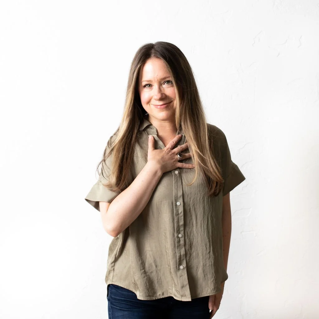 A woman with long brown hair, smiling and holding her hand on her chest, standing against a white background.