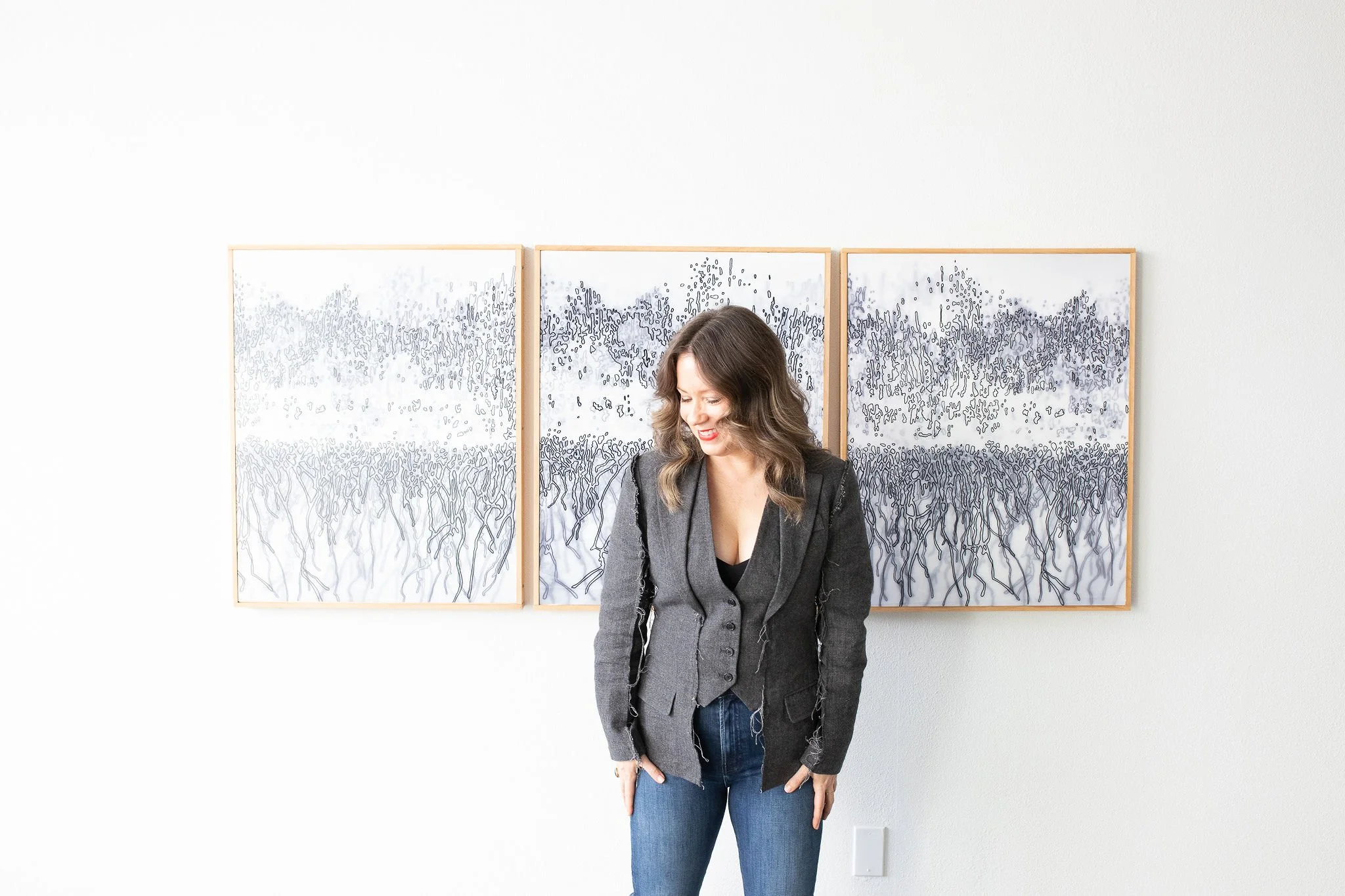 Woman with wavy brown hair smiling and looking down, standing in front of modern abstract black-and-white striped artwork on a white wall, wearing a gray blazer and jeans.