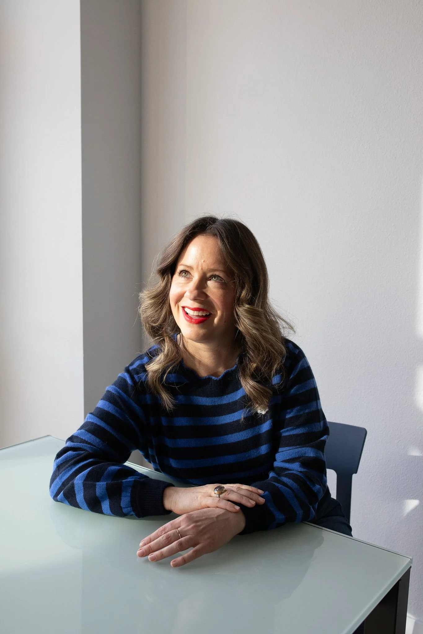 A woman with wavy brown hair and wearing a navy blue and black striped sweater, sitting at a white table, smiling and looking upward in a bright room with a white wall.