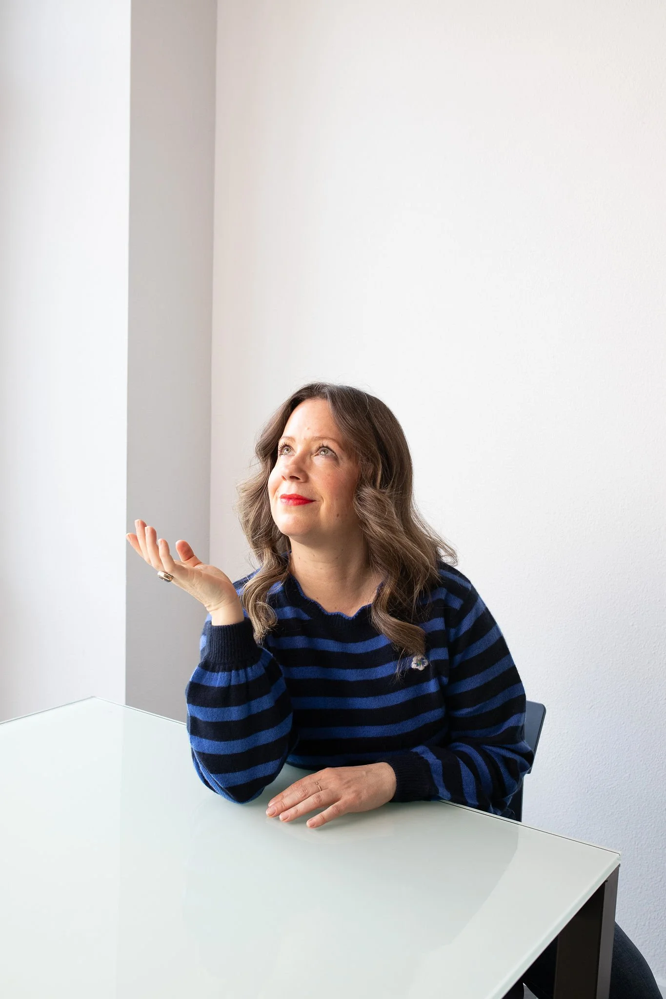 A woman with long, wavy brown hair and red lipstick sits at a white table in front of a mirror with white walls in the background. She is wearing a blue and black striped sweater and looking up while gesturing with her right hand.