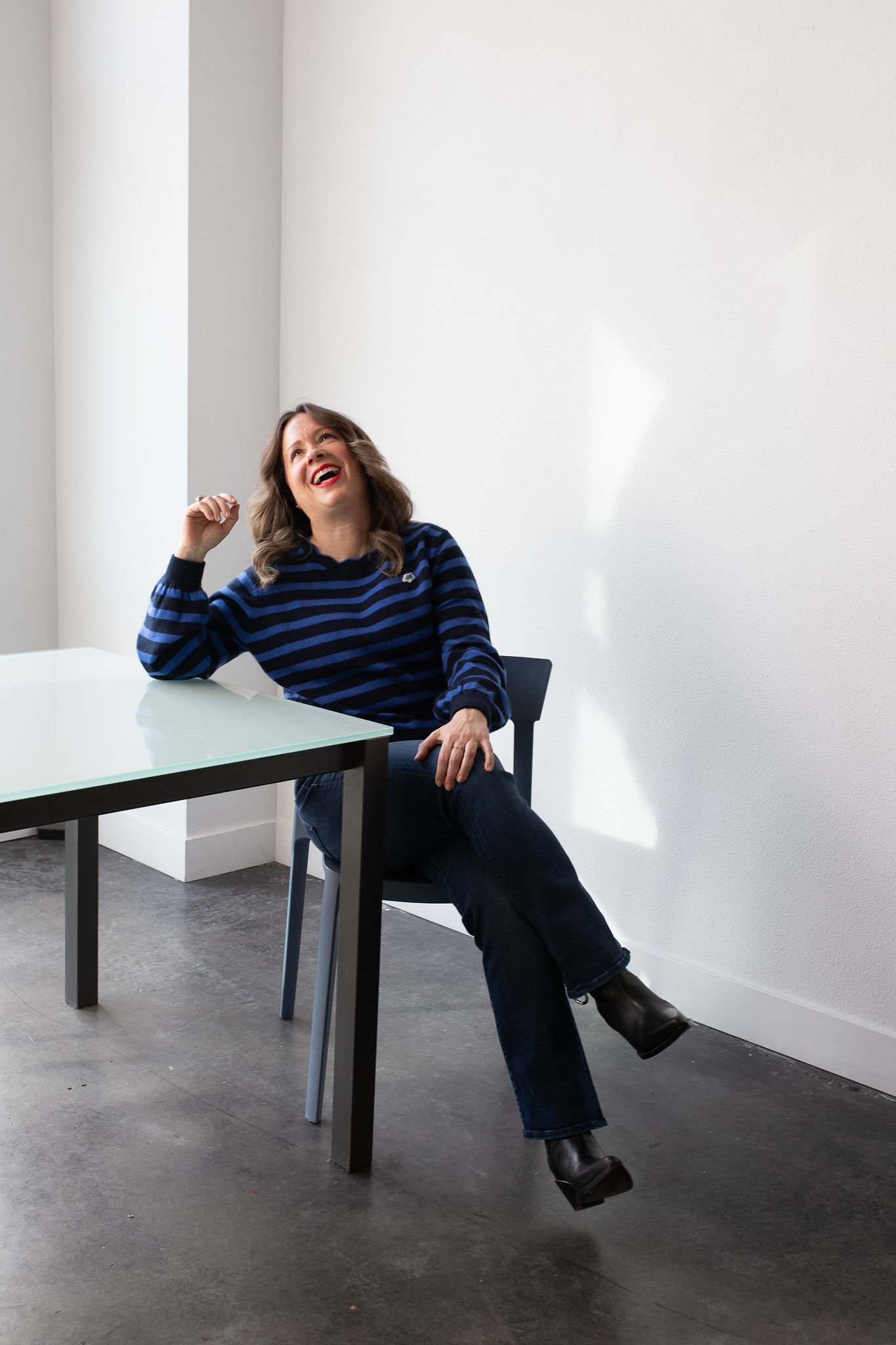 A woman with shoulder-length wavy hair, wearing a blue and black striped sweater, dark jeans, and black heeled boots, sitting at a table with a white surface, laughing and gesturing with her right hand. The background features a plain white wall and a gray floor.