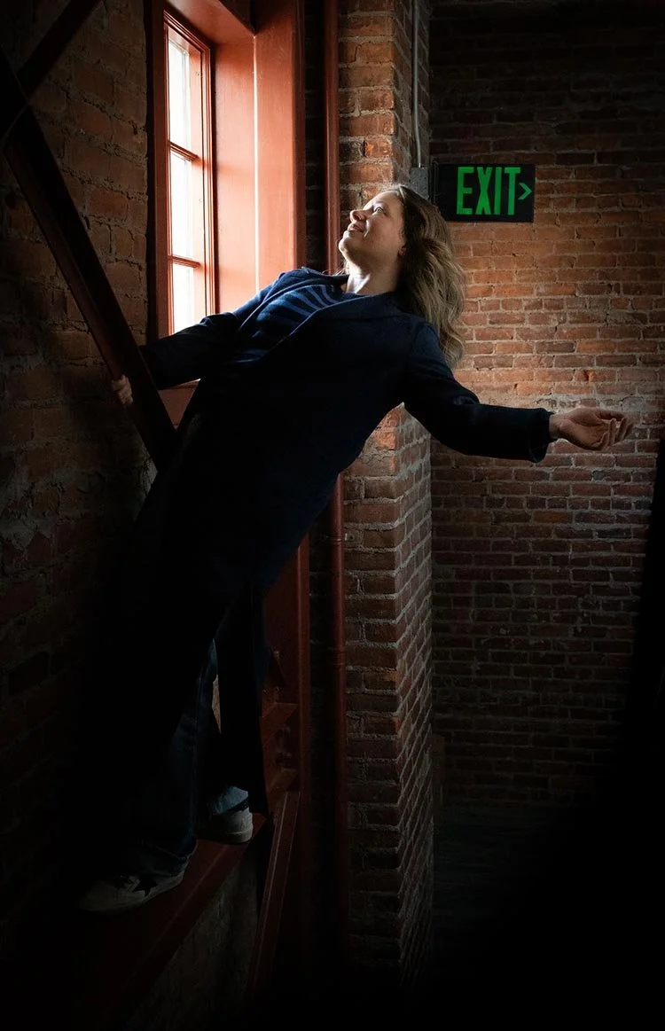 Person leaning against a brick wall next to a window, with arms outstretched and eyes closed, standing on a staircase near an exit sign.
