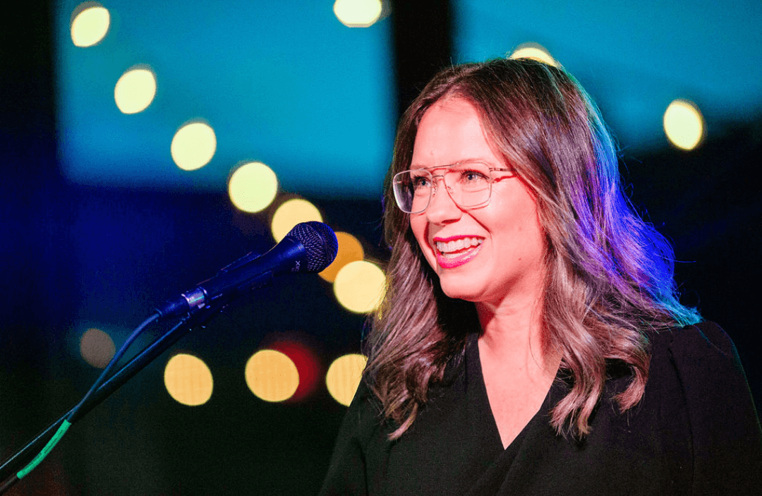 A woman with glasses and shoulder-length hair smiling while speaking into a microphone at an event with blurred colorful background lights.