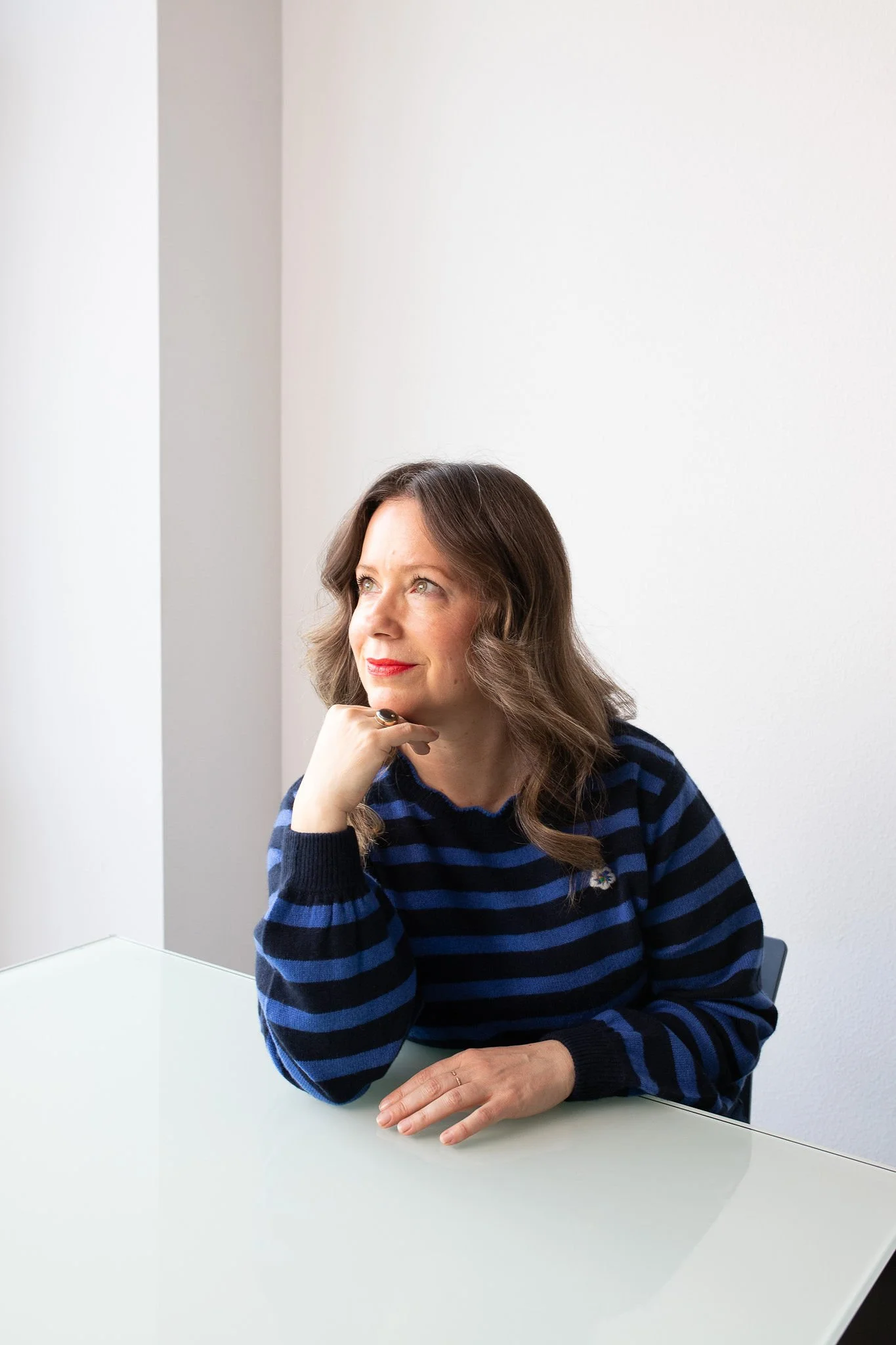 A woman with brown hair, wearing a blue and black striped sweater, sitting at a white table, resting her chin on her right hand, looking thoughtfully to her left in front of a plain white wall.