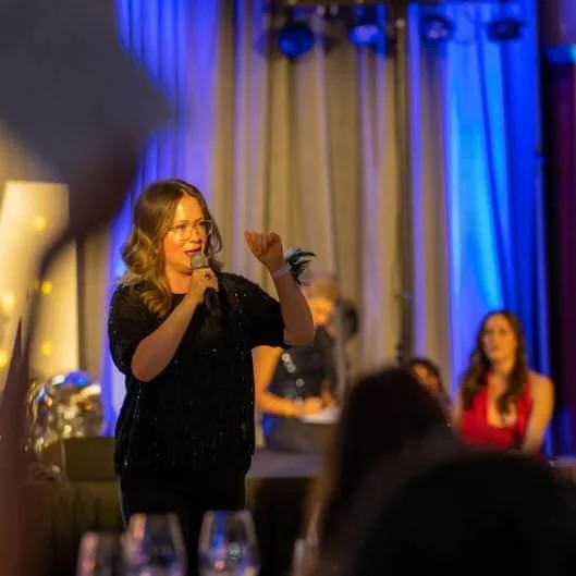 A woman with glasses and wavy hair speaking into a microphone at an event, with a blue and white curtain backdrop and seated audience members.