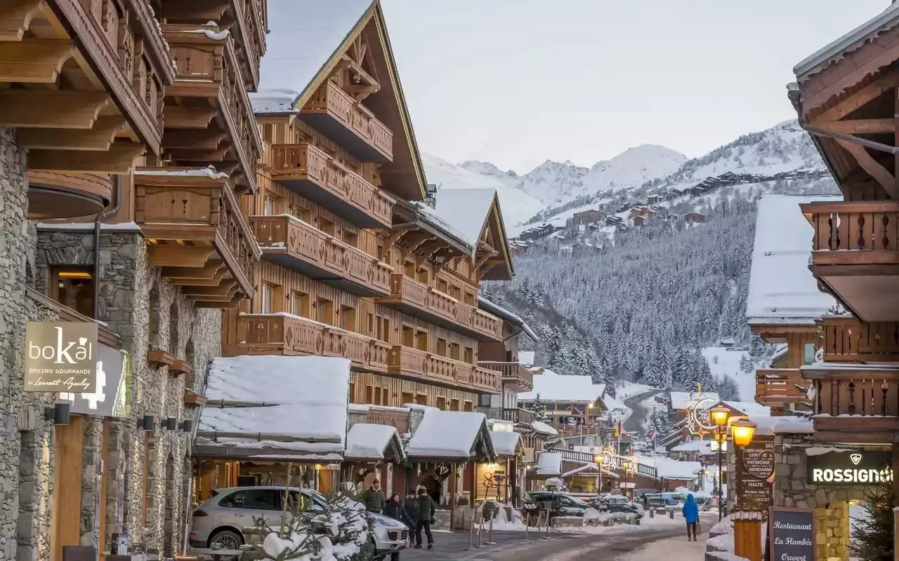 Exterior view of Hôtel La Chaudanne 4* in Méribel at dusk. The hotel, a large traditional-style chalet in stone and wood, is snow-covered and illuminated, creating a welcoming atmosphere.