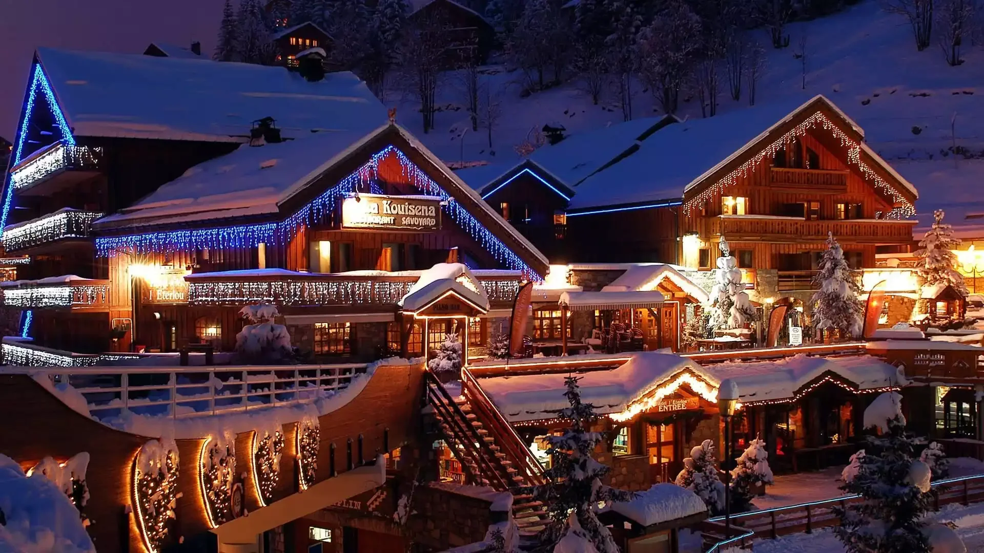 Night view of Hôtel L'Eternlou in Méribel. The wooden chalets, covered in snow and decorated with festive lights, create a warm and welcoming atmosphere in the mountains.