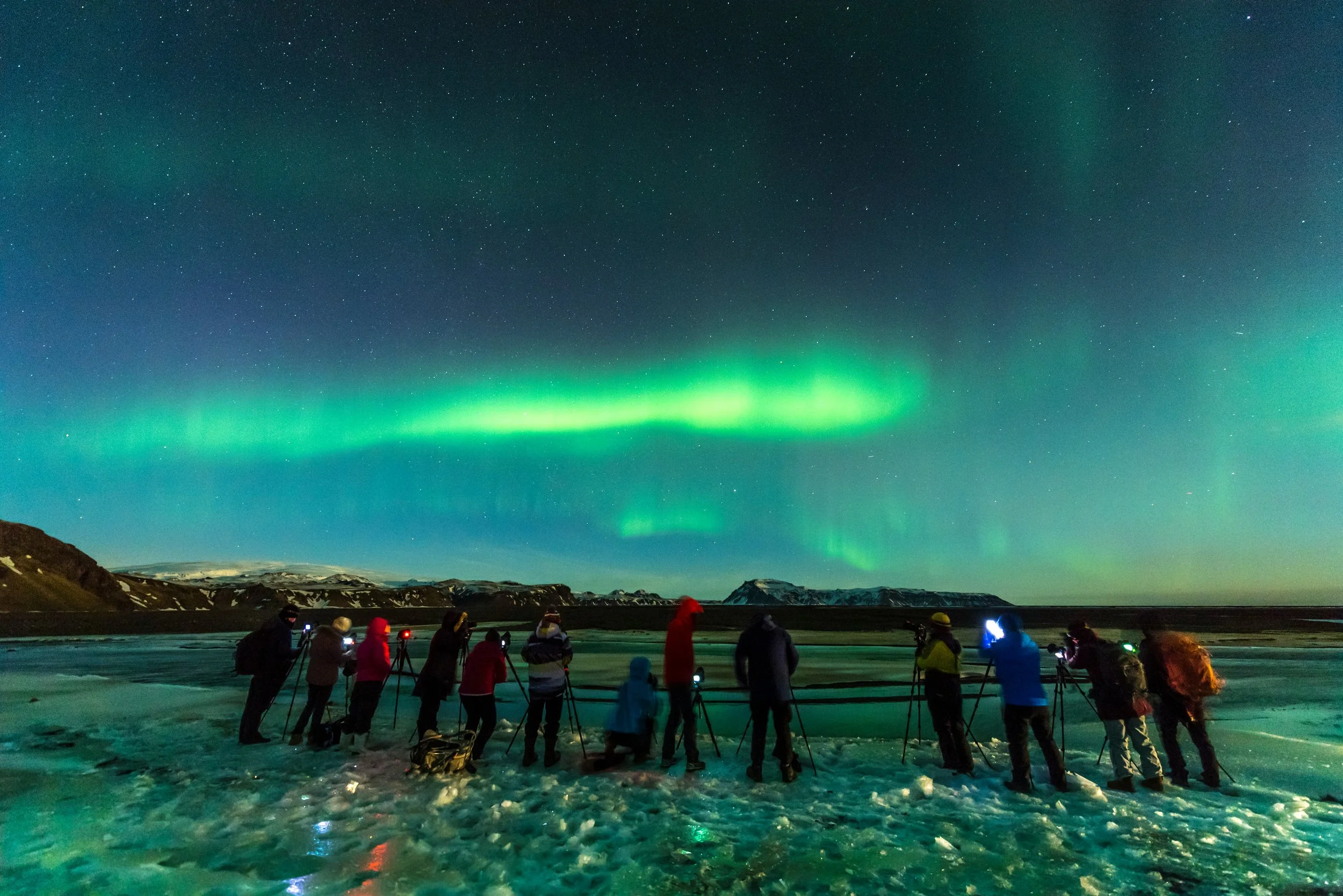 Spectacle d'aurores boréales vertes et brillantes au-dessus d'un paysage islandais. Un groupe de personnes utilise des trépieds pour photographier le phénomène, représentant une activité Aylo Montagne unique.
