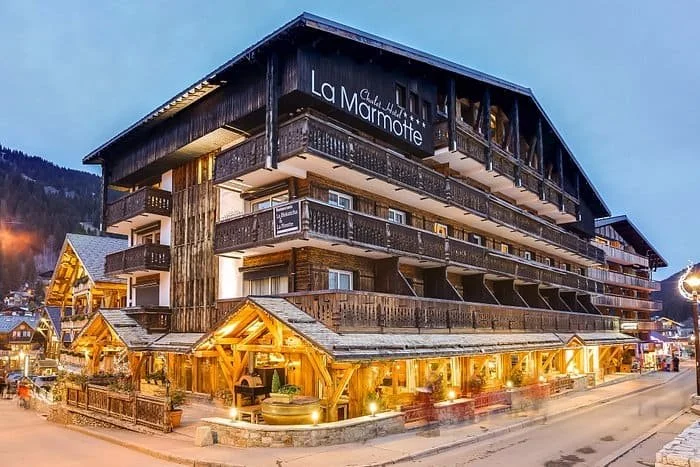 Façade illuminée de La Marmotte Hôtel la nuit. Ce grand chalet de bois traditionnel, couvert de neige, est un lieu d'accueil idéal pour un séminaire d'entreprise Aylo Montagne chaleureux.