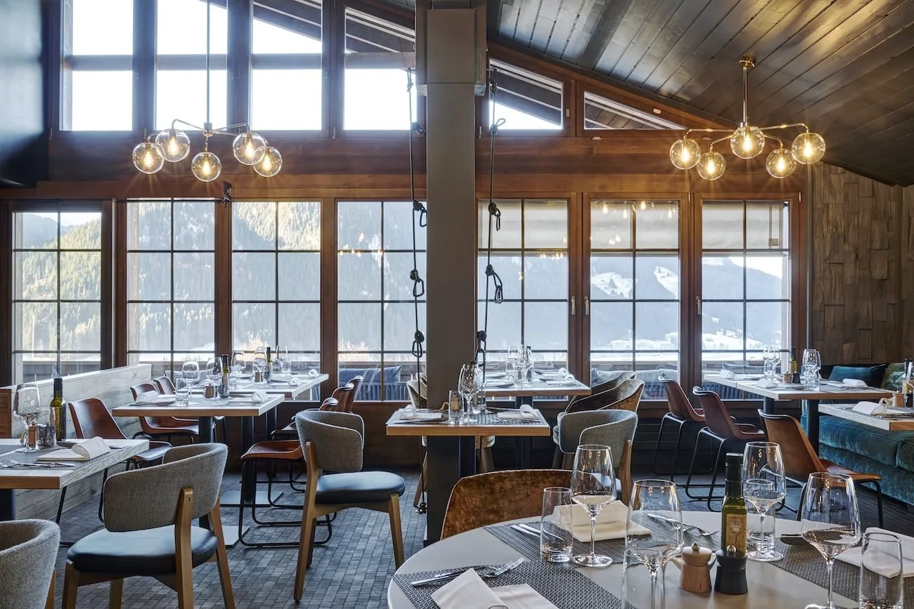 Restaurant lumineux de l’hôtel HUUS Gstaad, avec grandes baies vitrées, décoration contemporaine et tables élégamment dressées – Aylo Montagne.