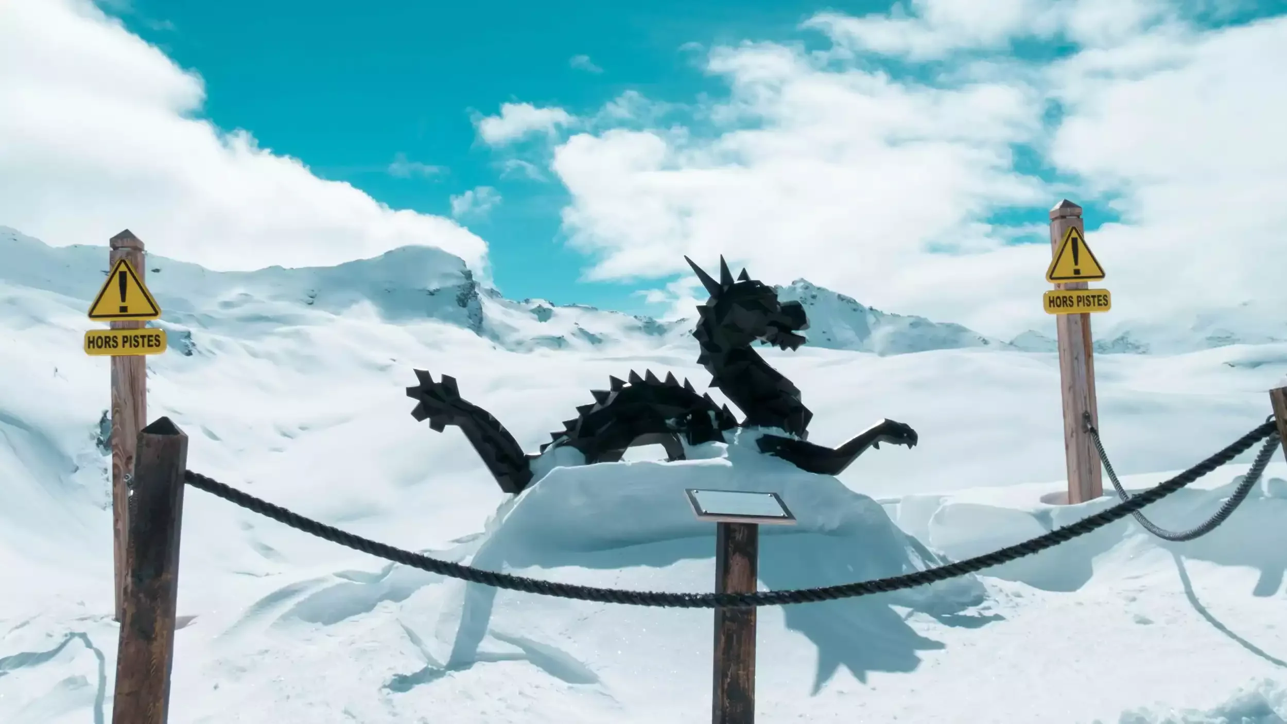 Sculpture of a black metal dragon emerging from the snow in Val d'Isère. The sculpture stands in a belvedere overlooking snow-capped mountains and a blue sky with scattered clouds.