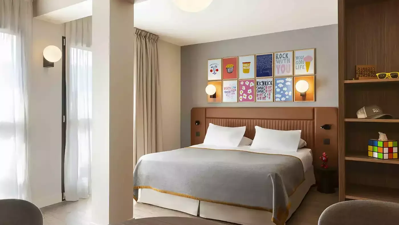 Chambre au design minimaliste et moderne à l’hôtel Rocky Pop. Le décor comprend des tableaux colorés sur un grand lit avec tête de lit en bois, créant un style pop art.