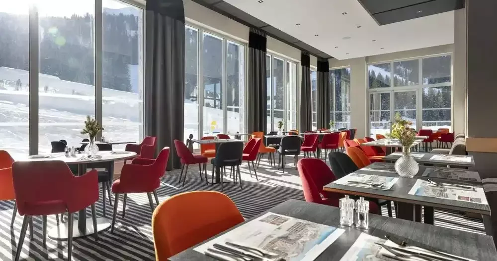 Restaurant de l’Alhena & spa hôtel avec de grandes baies vitrées offrant une vue directe sur les pistes de ski. Des tables rondes et des chaises aux couleurs vives créent une ambiance chaleureuse et moderne.
