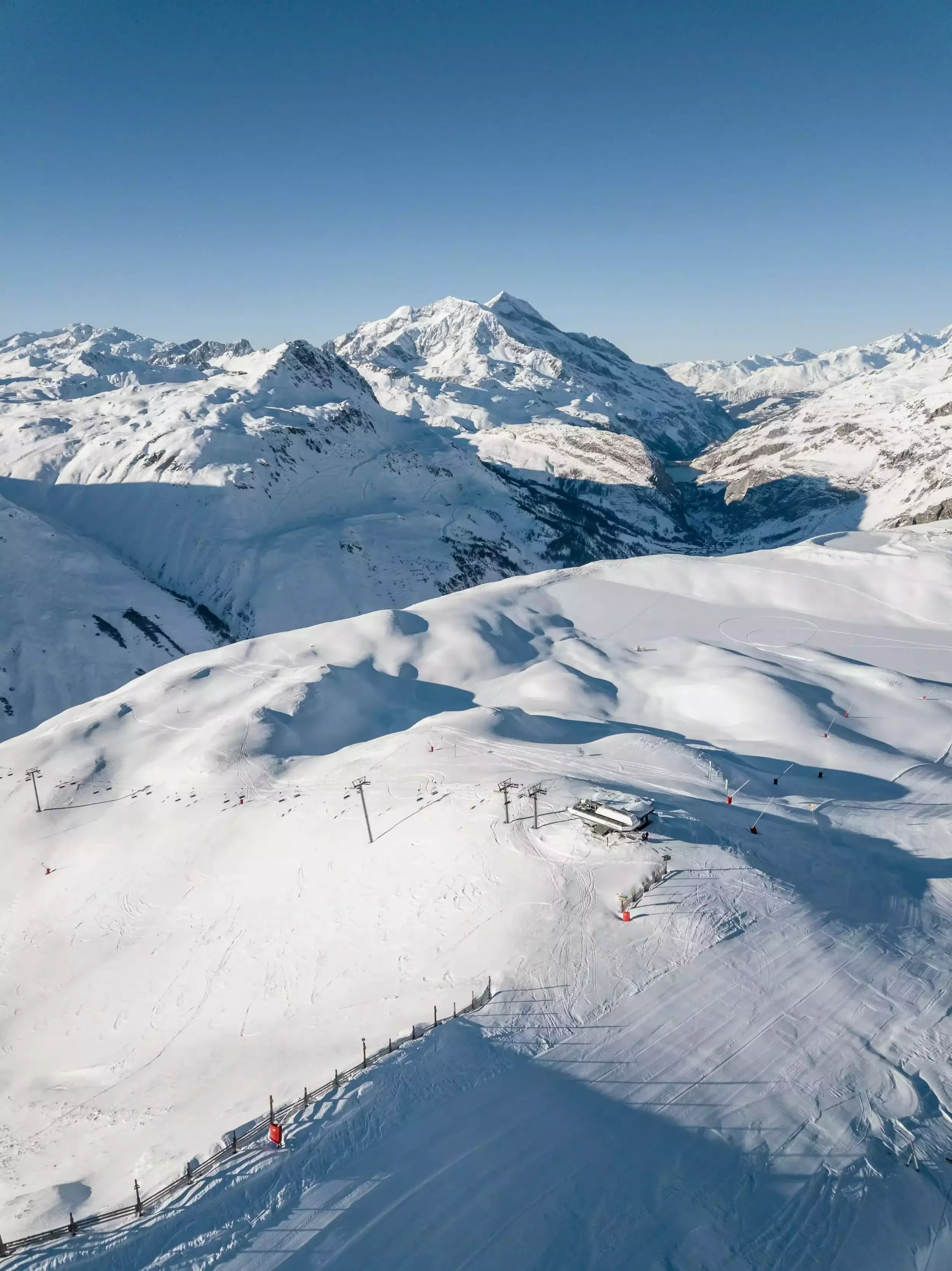 Aerial view of the snow-covered slopes of Tignes, with chairlift and mountain restaurant. In the background, snow-capped peaks rise beneath a brilliant blue sky.