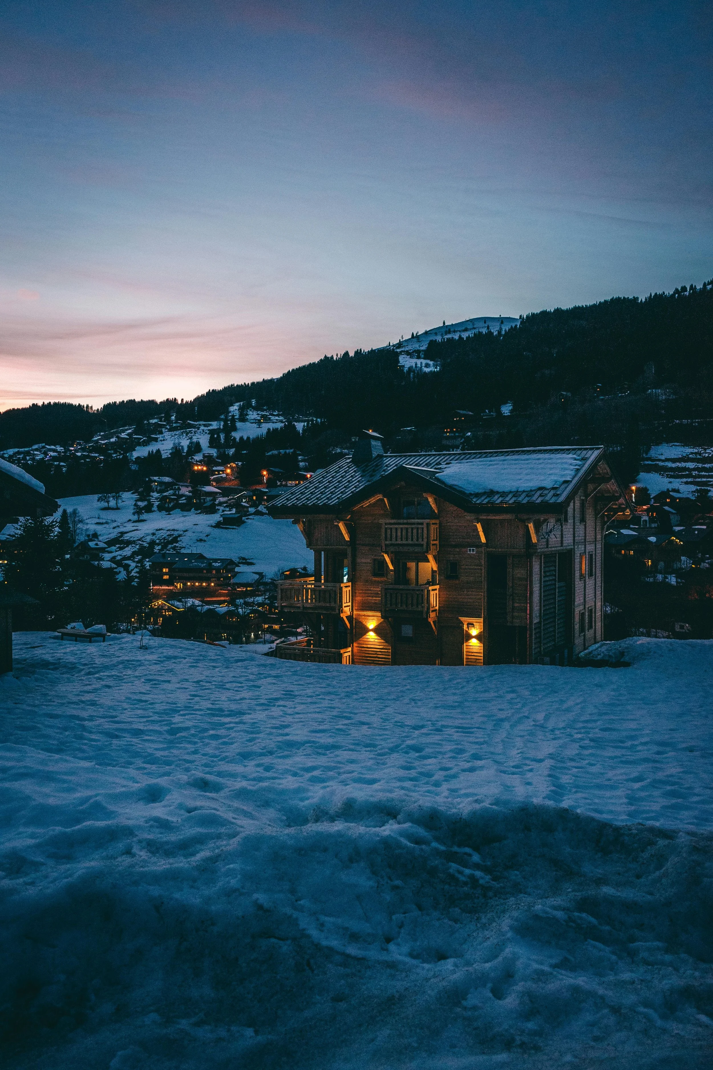 Chalet de bois isolé et illuminé dans la neige aux Gets, au crépuscule. Le cadre alpin est chaleureux et représente un hébergement idéal pour un séminaire d'entreprise Aylo Montagne.