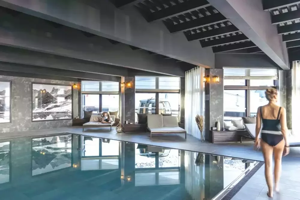 Indoor pool at Belambra clubs, with large windows offering a view of the snow-capped mountains. Sun loungers surround the pool, creating an atmosphere of tranquility and well-being.