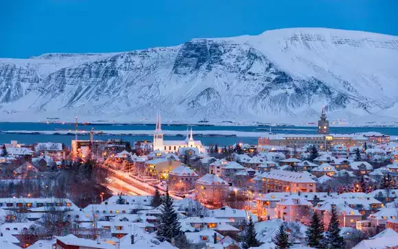 Vue panoramique de Reykjavik, Islande, au crépuscule. Les maisons de la ville sont couvertes de neige, avec des lumières chaudes contrastant avec les montagnes enneigées en arrière-plan. Un cadre idéal pour un séminaire Aylo Montagne.