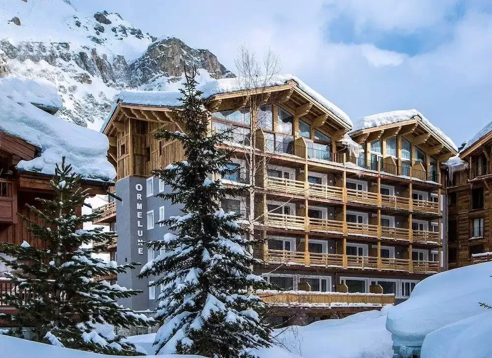 Facade of the Hotel Ormelune, a modern-style building combining a wooden structure with a grey concrete base. The hotel is covered in snow, with imposing mountains in the background.