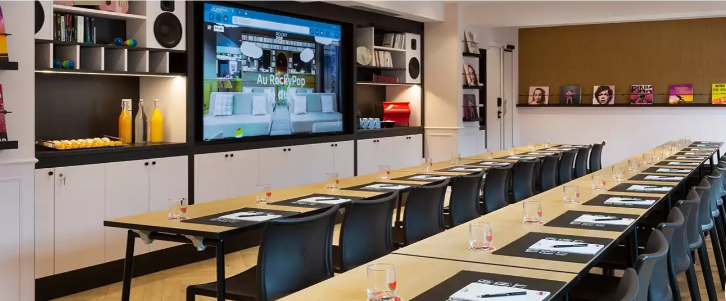 Salle de réunion à l’hôtel Rocky Pop, équipée d’une longue table et des chaises pour un séminaire d’entreprise. L’espace dispose d’un grand écran pour les présentations et des étagères avec des boissons et des collations.