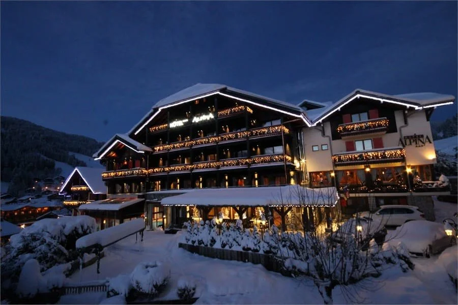 Façade extérieure de l'Hôtel Alpina aux Gets, un grand chalet de bois illuminé par des lumières de Noël. L'hôtel est couvert de neige, créant une ambiance festive pour un séminaire Aylo Montagne.