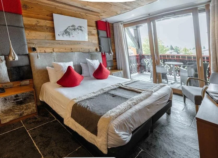 Chambre de style chalet à l'Hôtel Alpina Les Gets, avec murs en bois, grand lit et oreillers rouges. Le design est moderne et confortable, idéal pour le repos durant un séminaire Aylo Montagne.