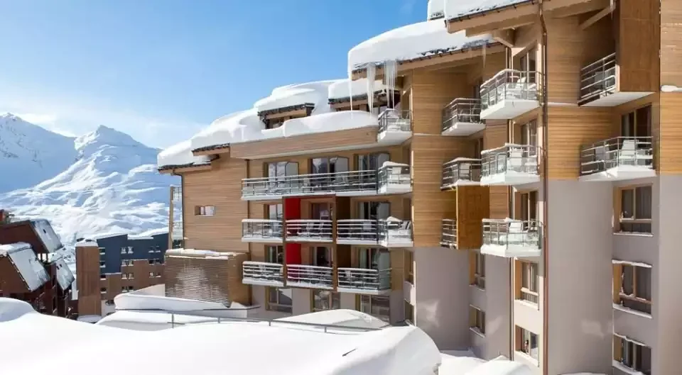Outdoor view of the Club Med Val Thorens Sensations on a sunny winter day. The hotel, covered in snow and ice, has a modern design with a wooden facade and red details on the balconies.