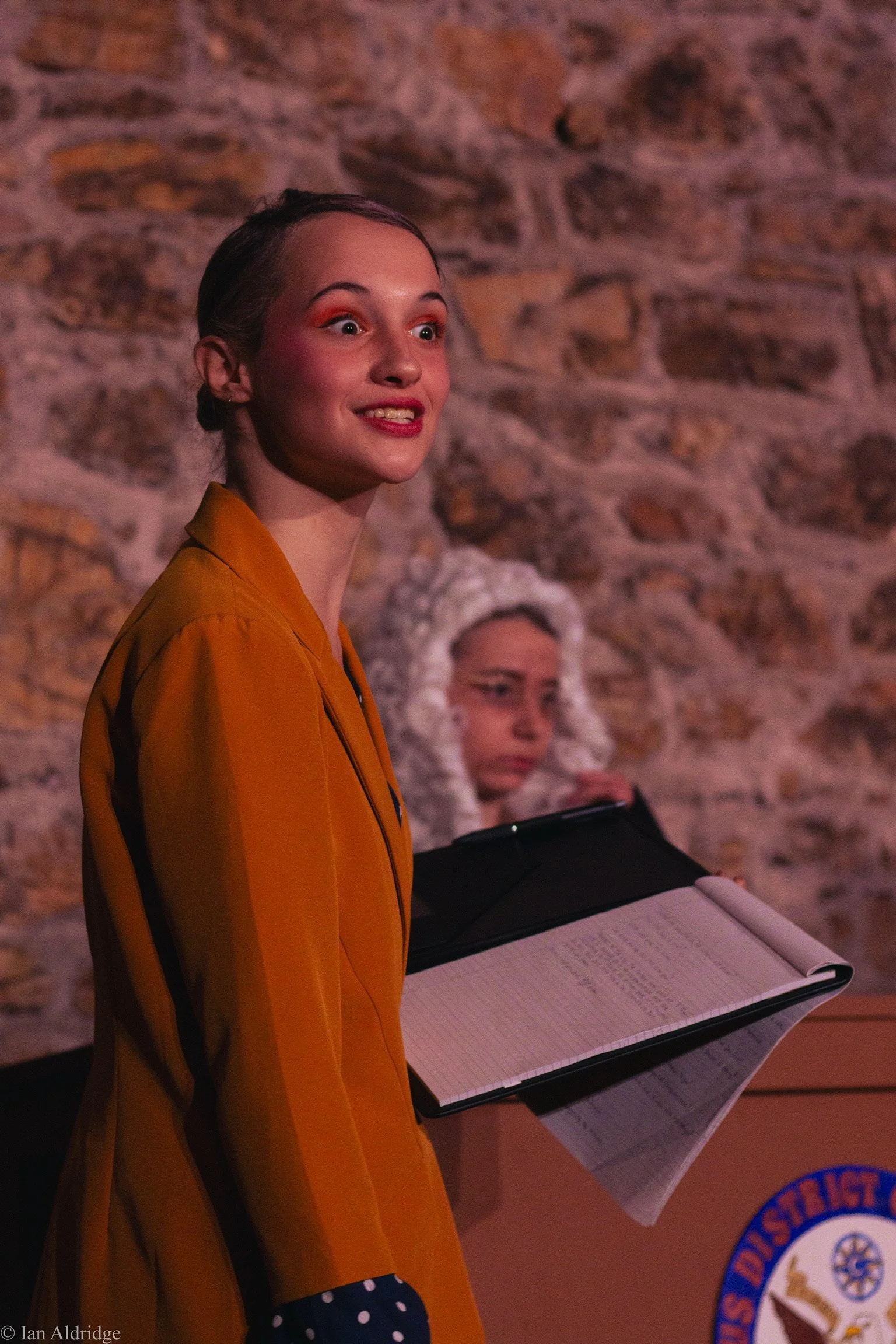 A young woman with a bright smile, wearing an orange jacket, holding a black folder or notebook, standing against a brick wall. Behind her, there is another woman with white hair wearing a beige hoodie and black cap, sitting at a table with some docu