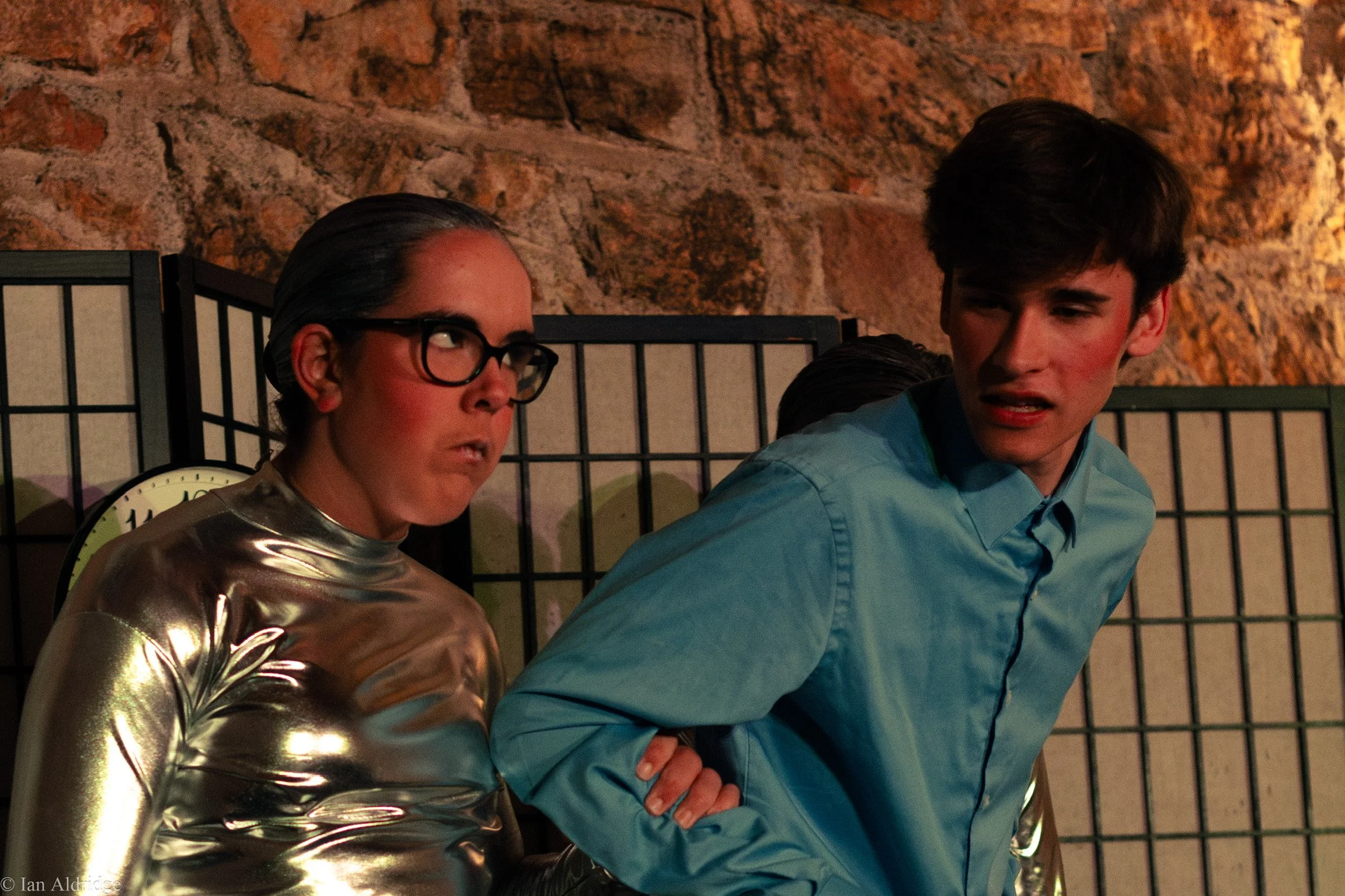 A woman with glasses and a metallic gold top sitting next to a young man with dark hair wearing a blue shirt, both seated in front of a stone wall and a black decorative divider. The woman is looking and speaking to the young man, who has his arms cr