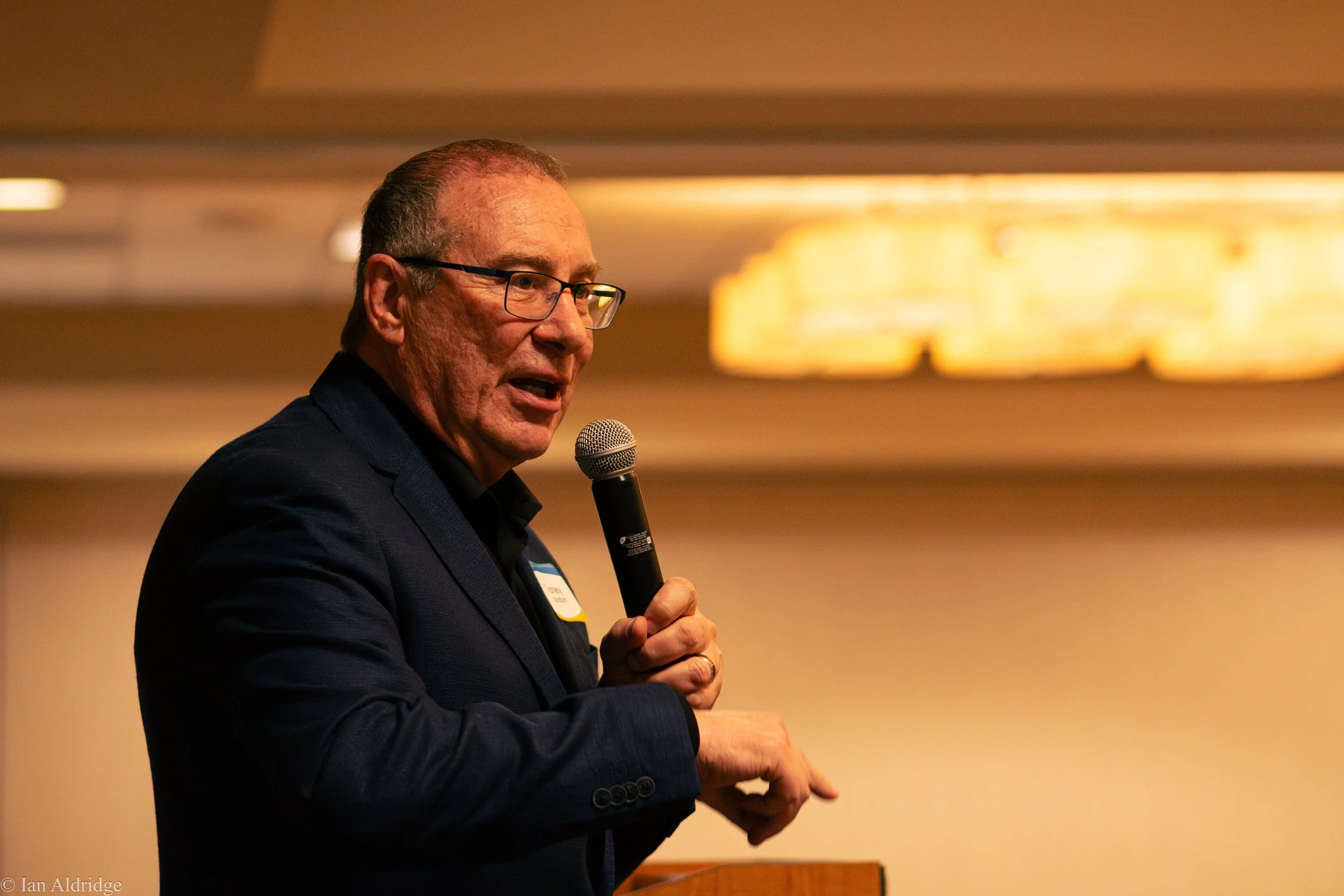 A man in glasses and a dark suit speaking into a microphone at an indoor event.