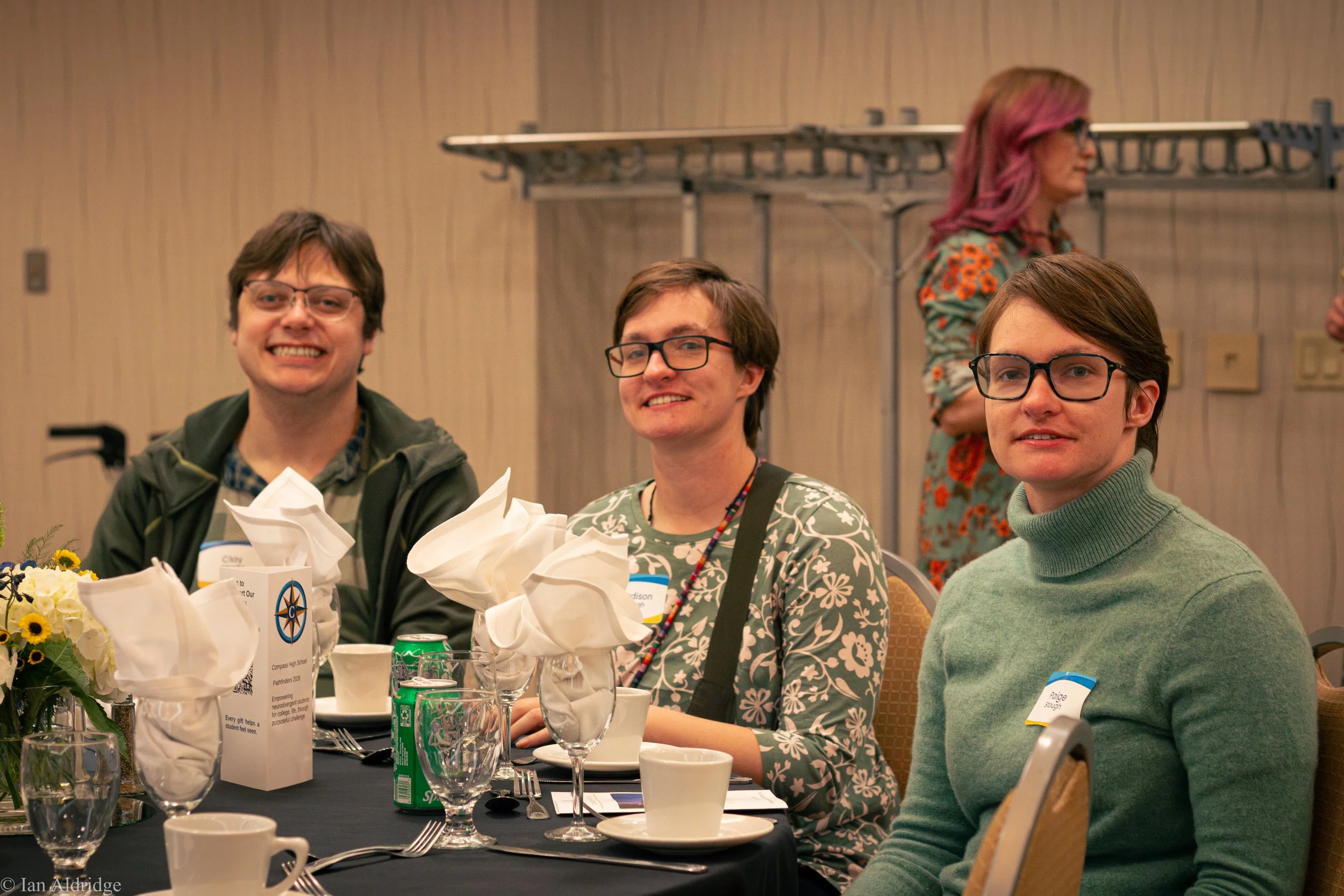 A group of four people sitting at a conference table at a formal event, with drinks, flowers, and name tags in front of them. Three men are in the foreground, smiling and looking at the camera, while a woman with pink hair and glasses is in the backg