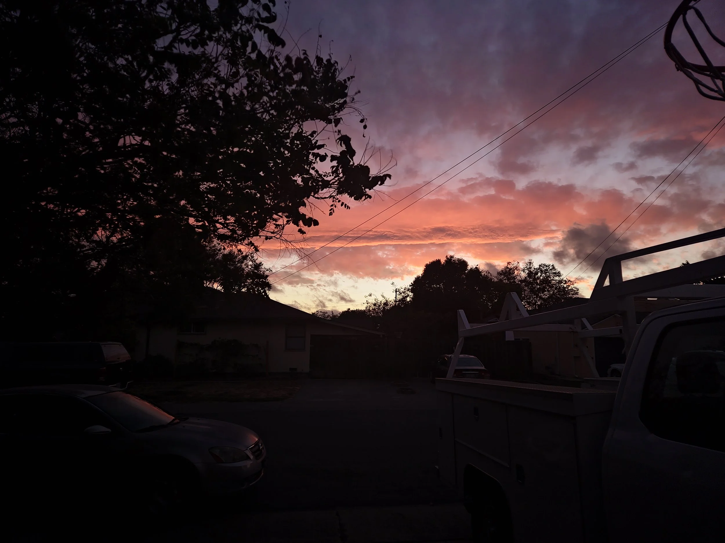 Sunset sky with colorful clouds, silhouettes of trees and power lines, and parked cars in a residential area.