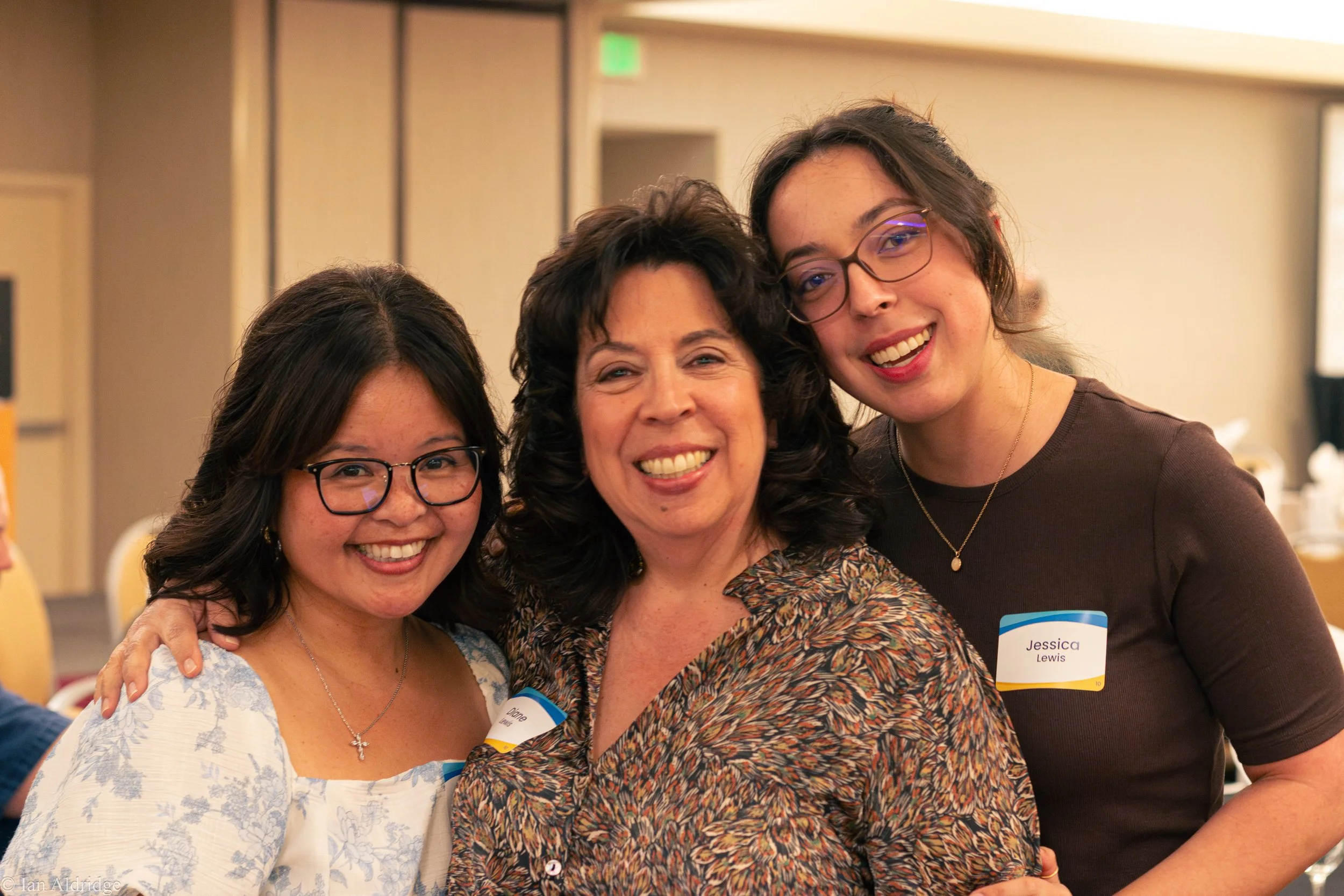 Three women smiling and posing for a photo, one wearing glasses, another with curly hair and a patterned blouse, the third with glasses and wearing a name tag that reads 'Jessica Lewis', in a conference or event setting.