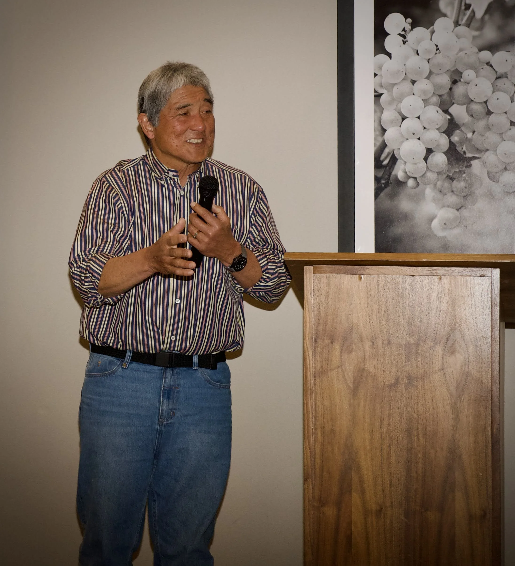 A man with gray hair wearing a striped shirt and blue jeans, holding a microphone, speaking or presenting in a room with a white wall and a large black-and-white photograph of grapes on displayed in the background.