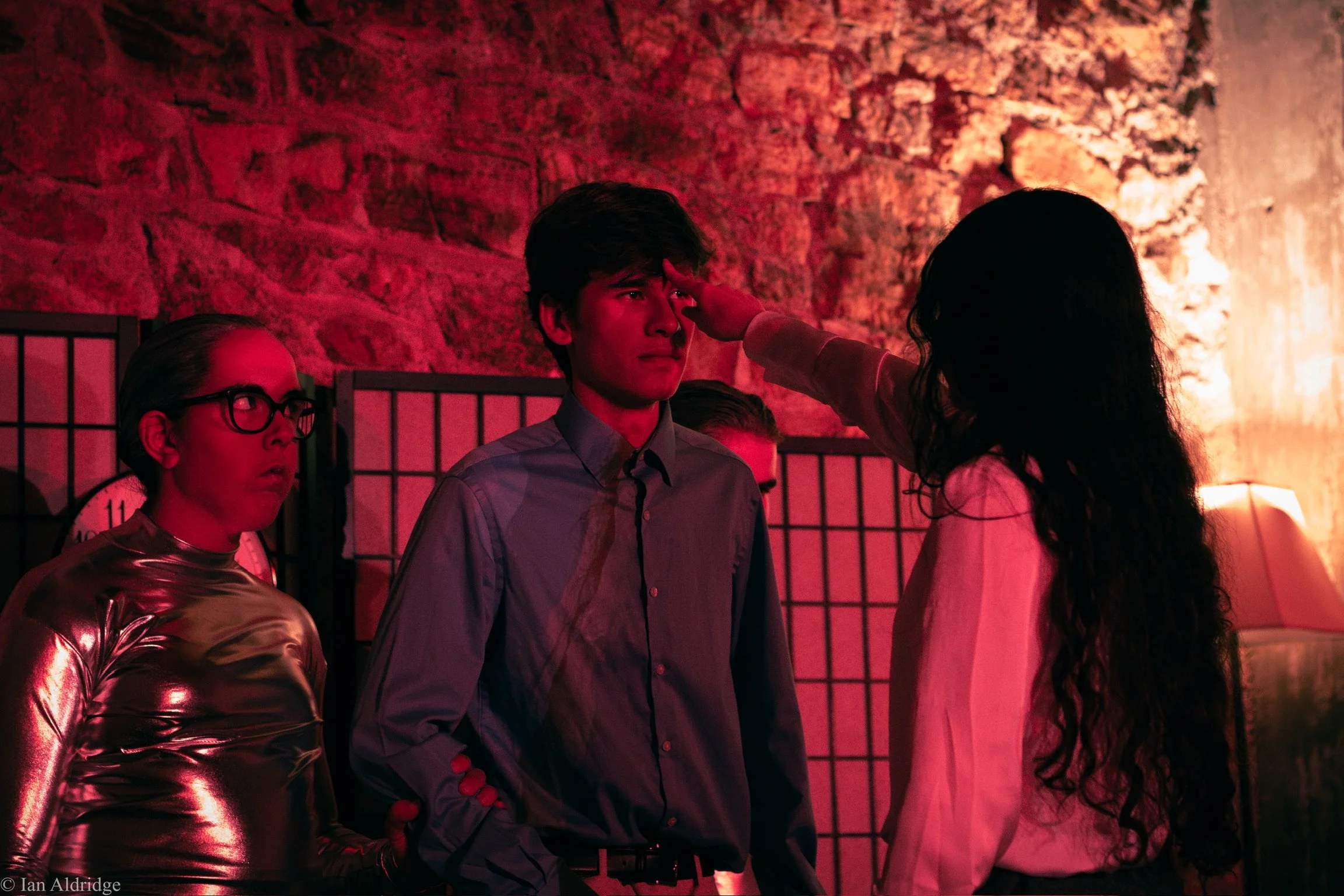 A young man is having his forehead touched or examined by a woman with long dark hair, in a dimly lit setting with red lighting and a stone wall background. Another woman with glasses and a shiny, metallic top observes nearby.