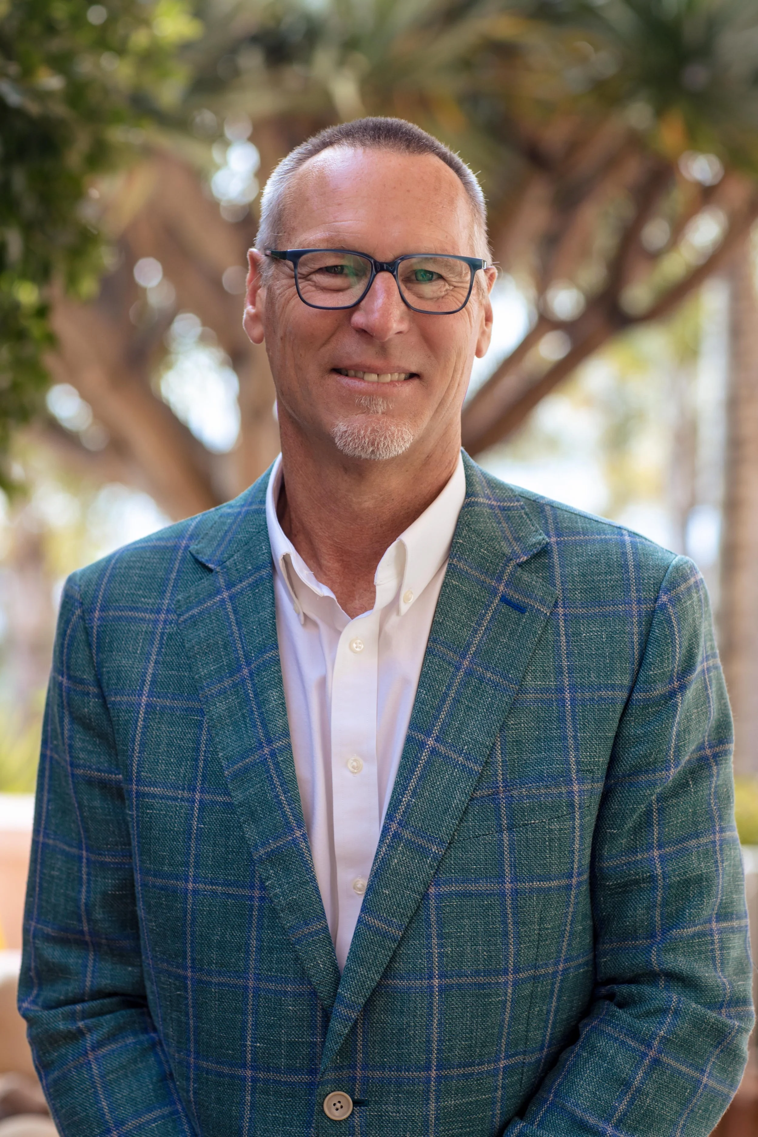 Portrait of a middle-aged man wearing glasses and a plaid blazer, smiling outdoors with trees and sunlight in the background.