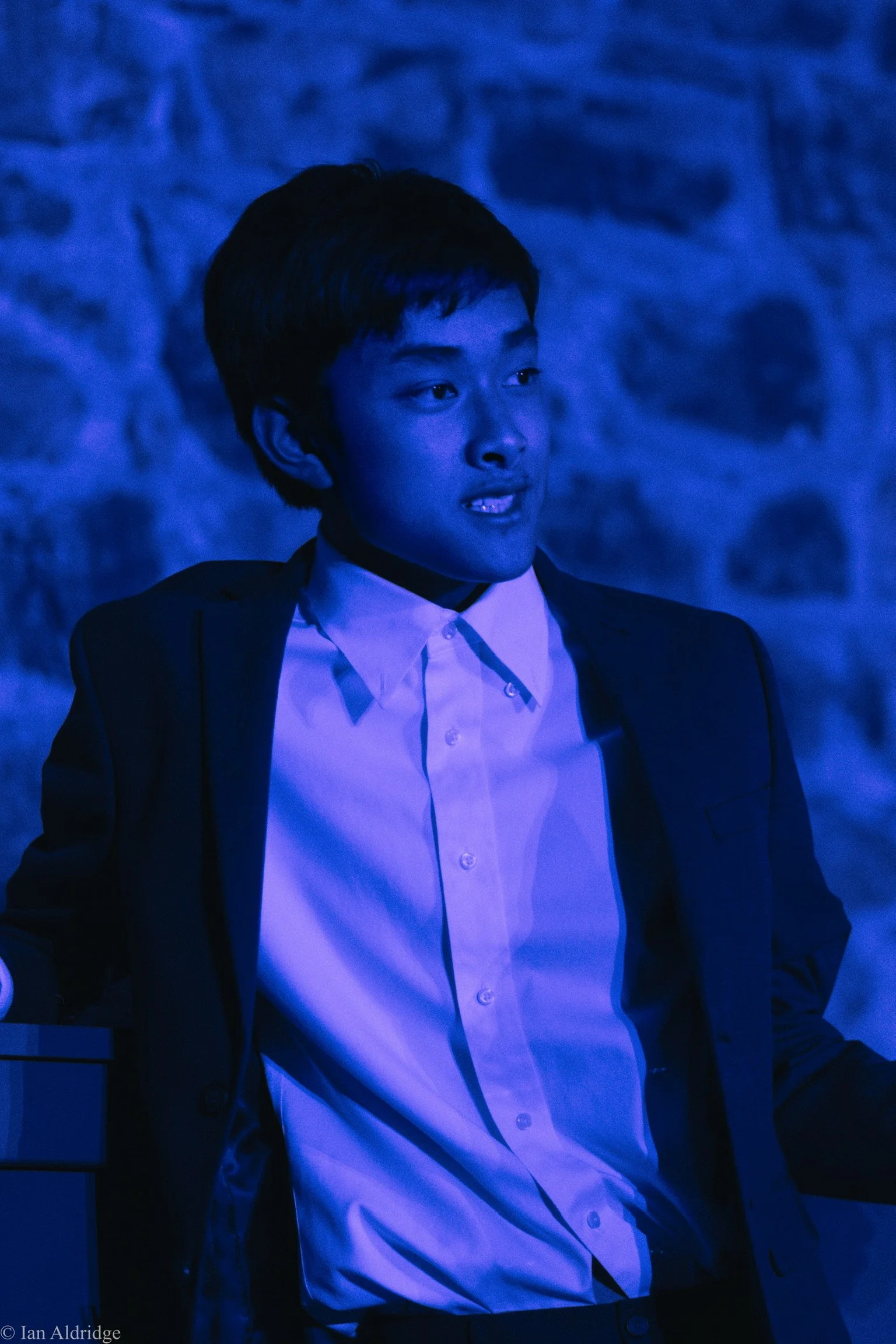 Young man in a suit with a white shirt, seated with a stone wall background, illuminated by blue light.
